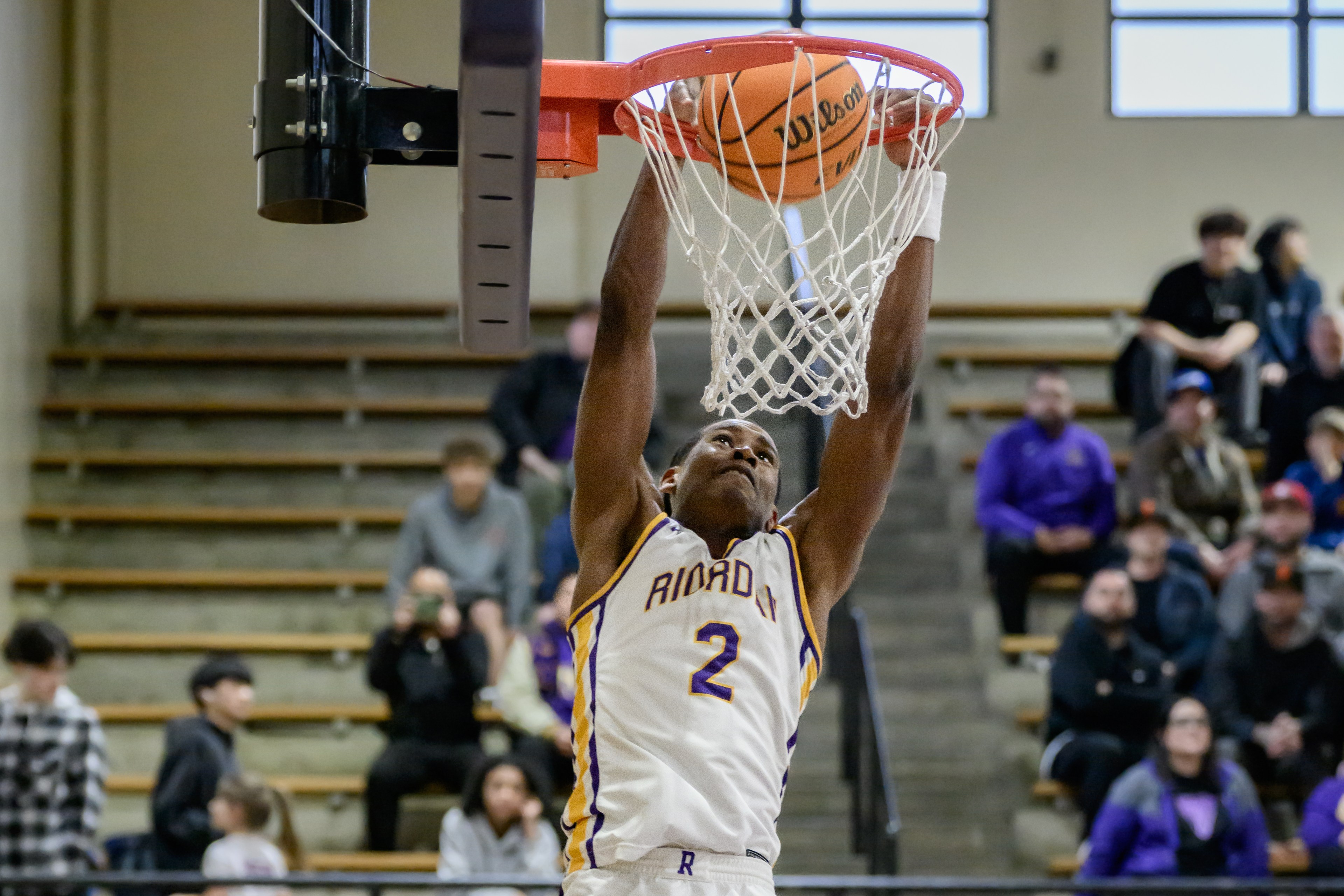 A basketball player is leaping to dunk a ball into the hoop. He's wearing a white jersey with purple and gold trim, and an audience watches from the background.