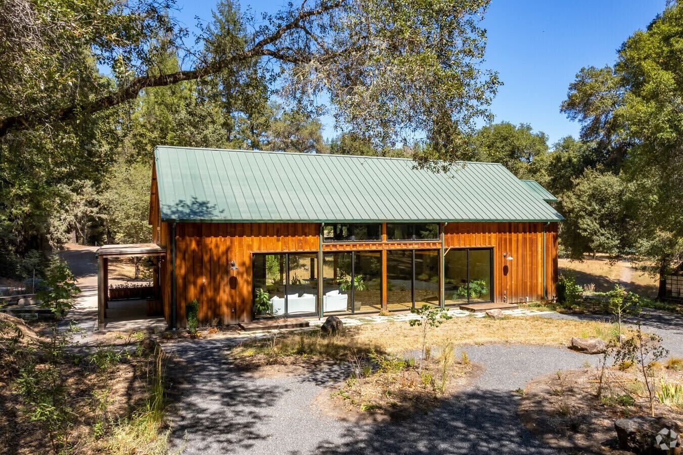 The image shows a wooden cabin with a green metal roof, surrounded by trees. Large glass windows are visible, and there's a gravel path leading to it.