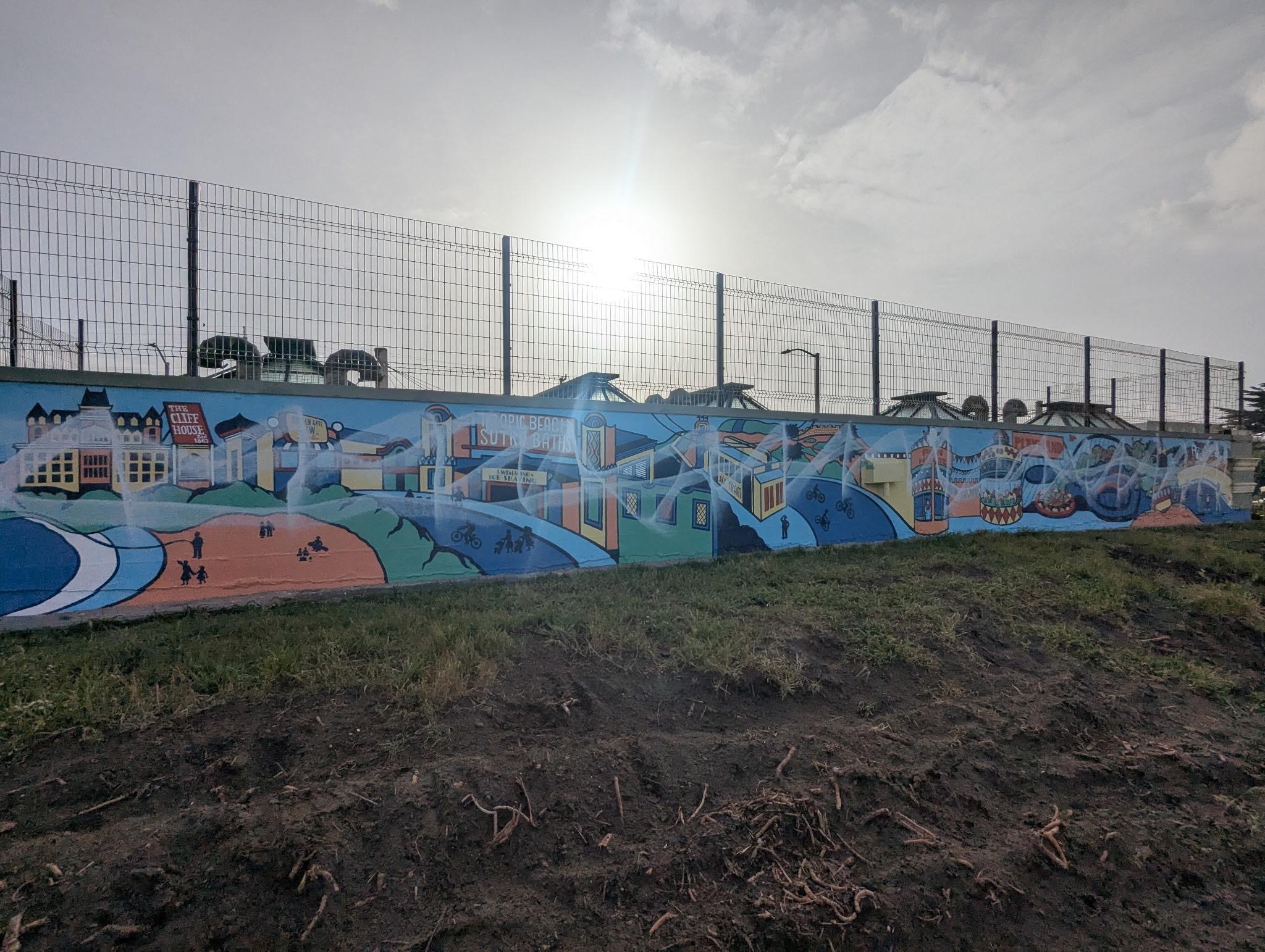 A vibrant mural features a scenic landscape with buildings, paths, and silhouettes of people. It spans a long wall under a bright, hazy sky.