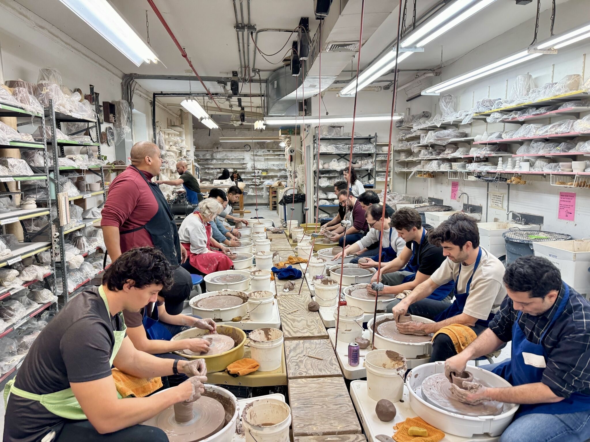 People are sitting in a row at pottery wheels, shaping clay. Shelves filled with supplies line the room, and a person is instructing the class.