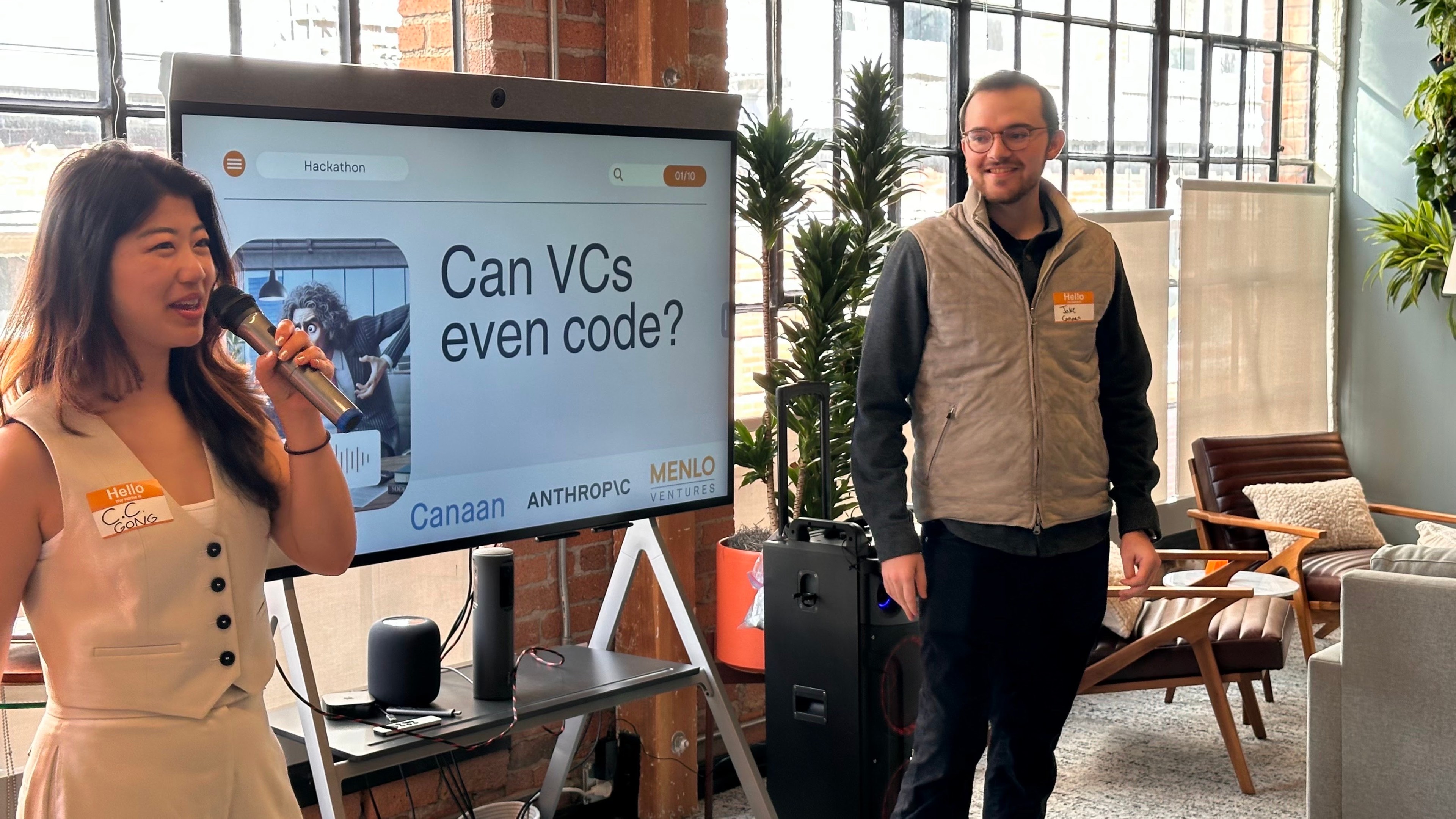 A woman with a microphone and a man stand near a screen displaying &quot;Can VCs even code?&quot; at a hackathon. The room has large windows and modern furniture.