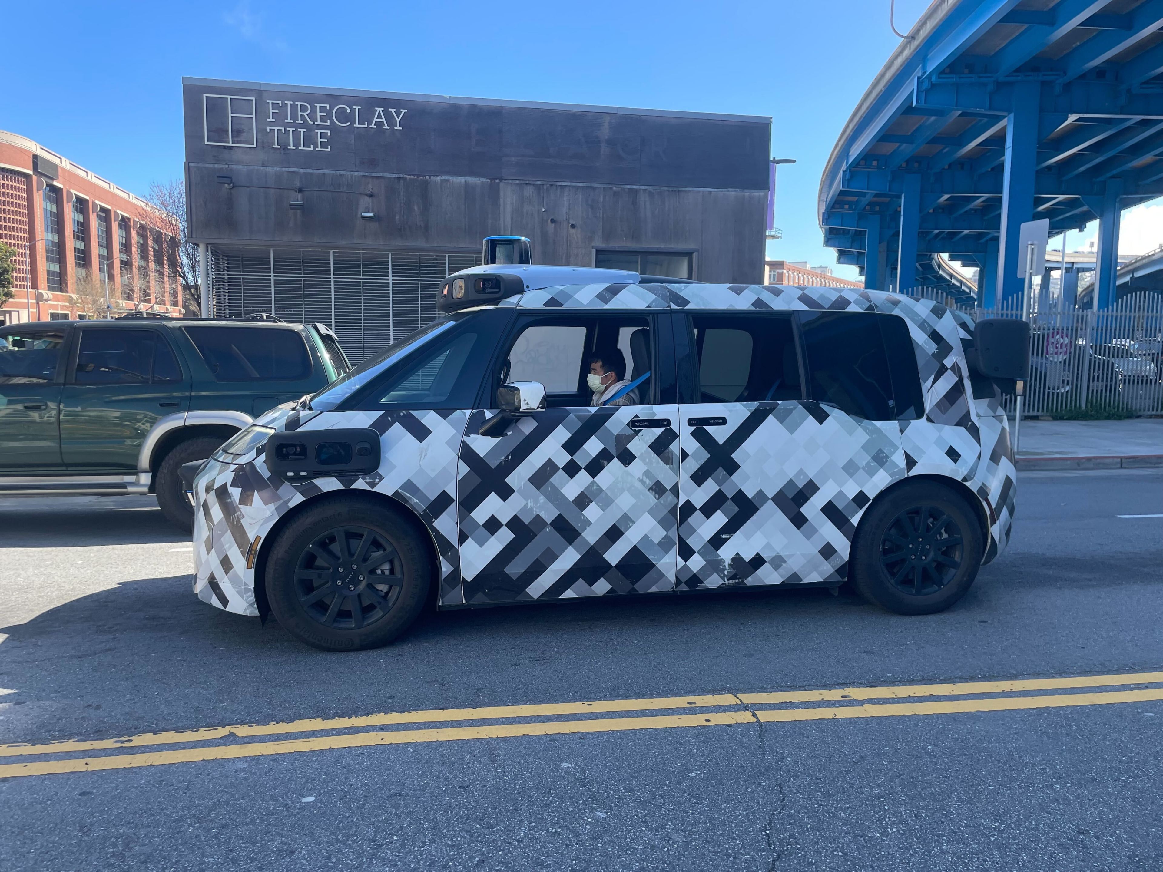 A camouflaged vehicle with a black-and-white pixelated pattern is parked on a street. A person inside is wearing a mask. A blue overpass and buildings are visible.