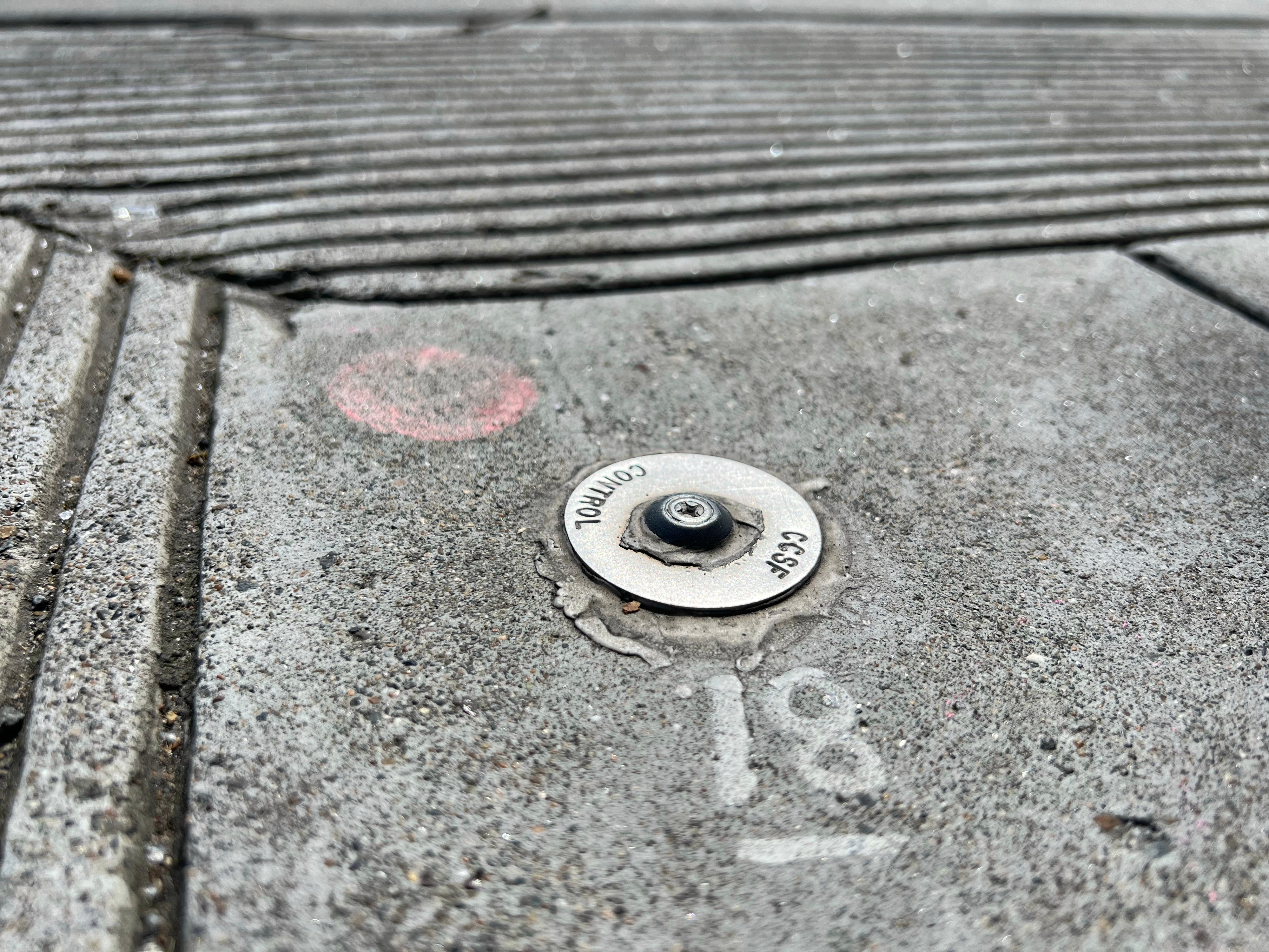 A concrete sidewalk with a textured grip pattern contains a small metallic disk labeled &quot;Control&quot; and &quot;CCSF,&quot; with faint numbers and a red circle nearby.