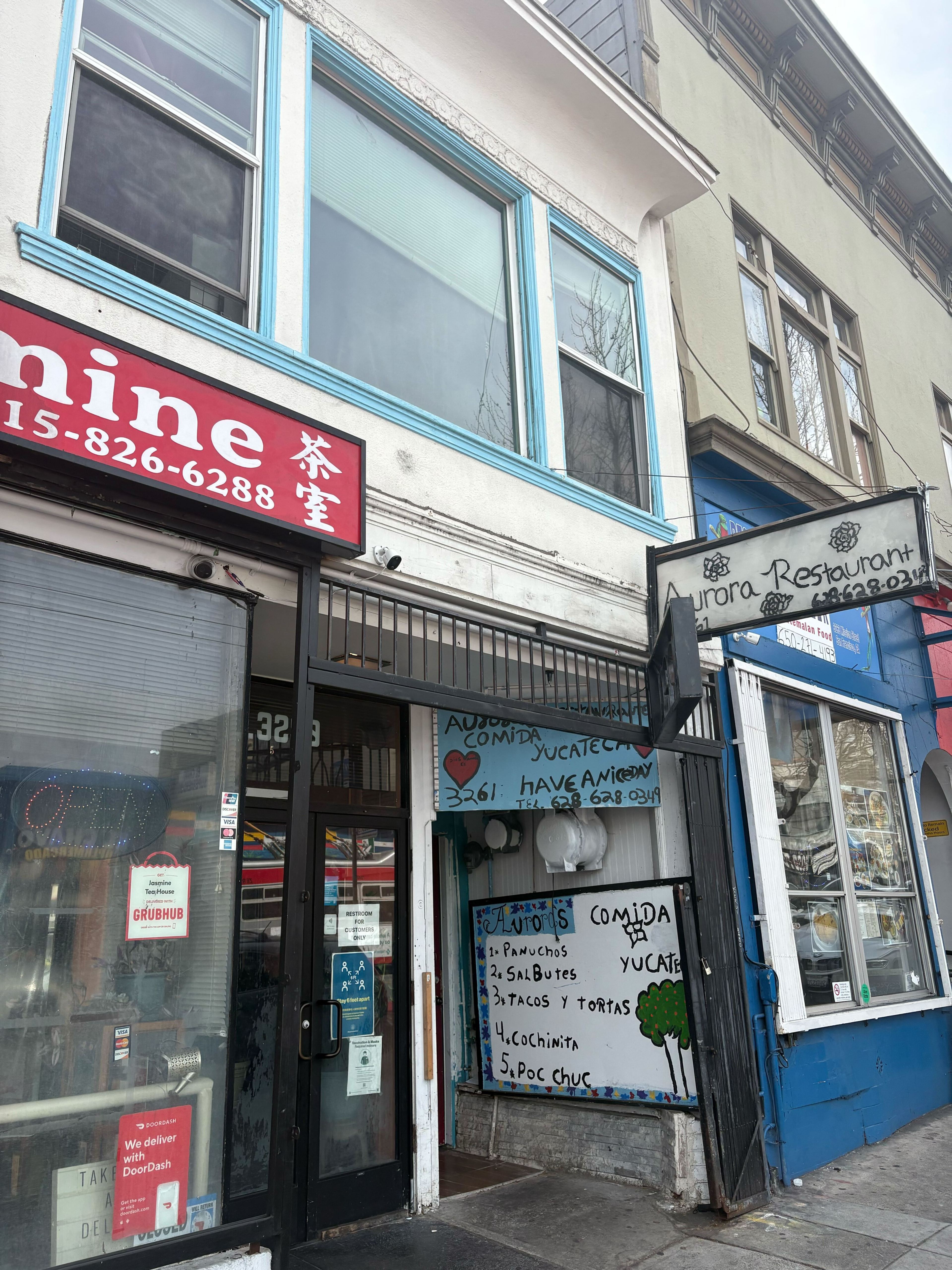 The image shows a street view featuring two buildings with signs for restaurants, including menu items on a board. The architecture has blue-framed windows and colorful signage.