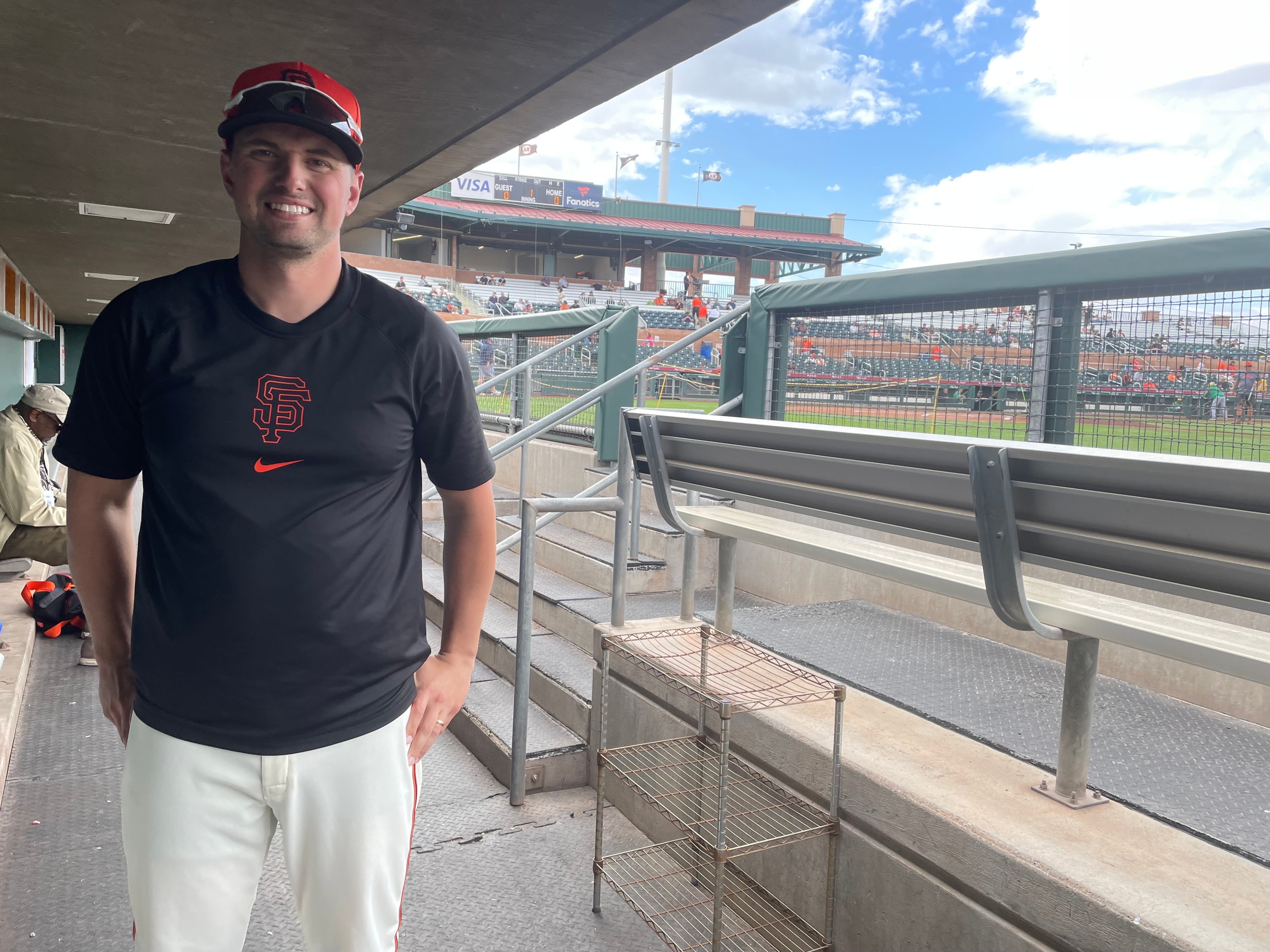 A person wearing a black San Francisco Giants shirt and cap stands in a baseball dugout, smiling. The field and stands with fans are visible behind them.