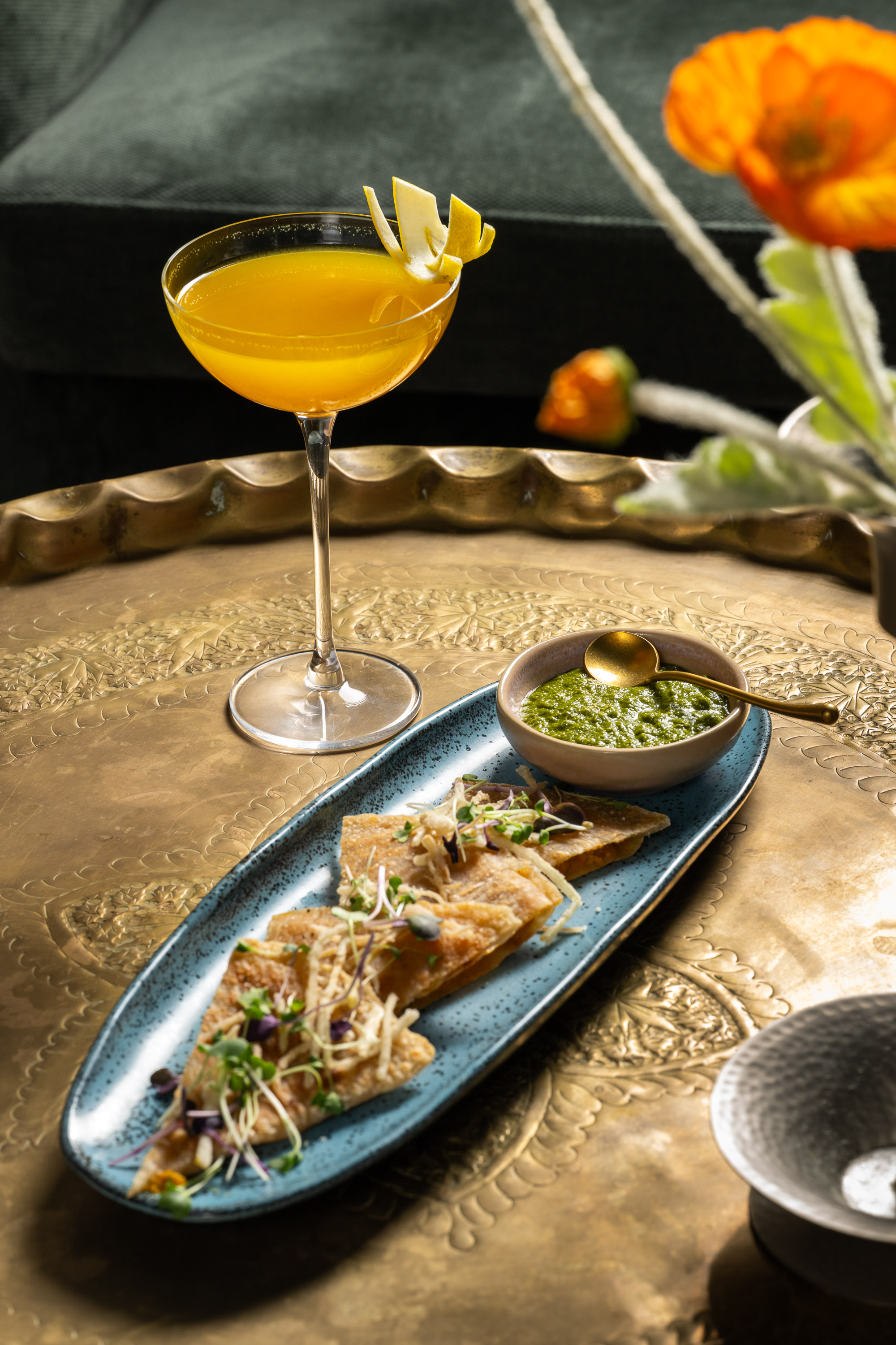 A golden cocktail with lemon garnish sits on a decorative brass table, alongside a blue plate of garnished triangular flatbreads and a bowl of green dip.