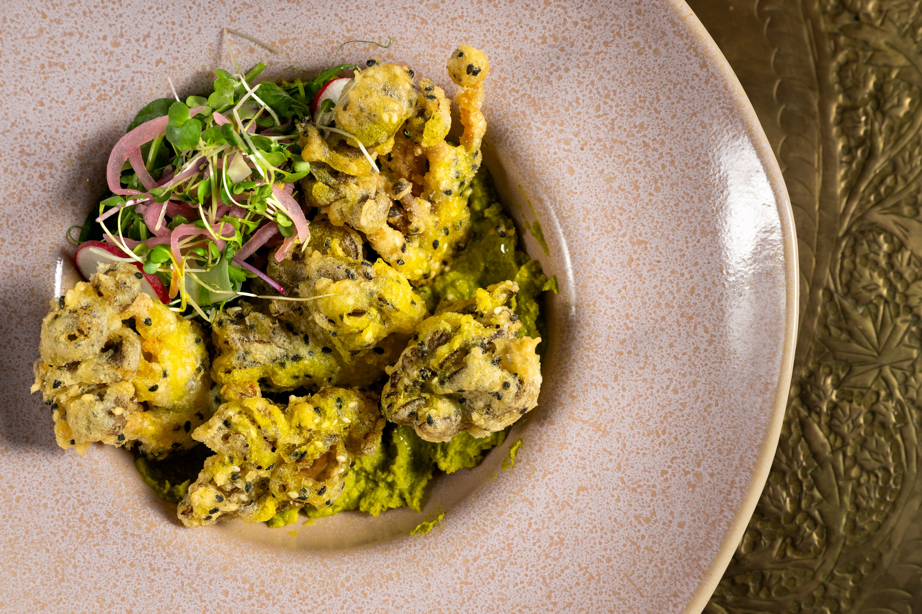 The image shows crispy, golden-brown fried fritters on a plate, accompanied by vibrant green sauce and garnished with fresh herbs and pink pickled onions.