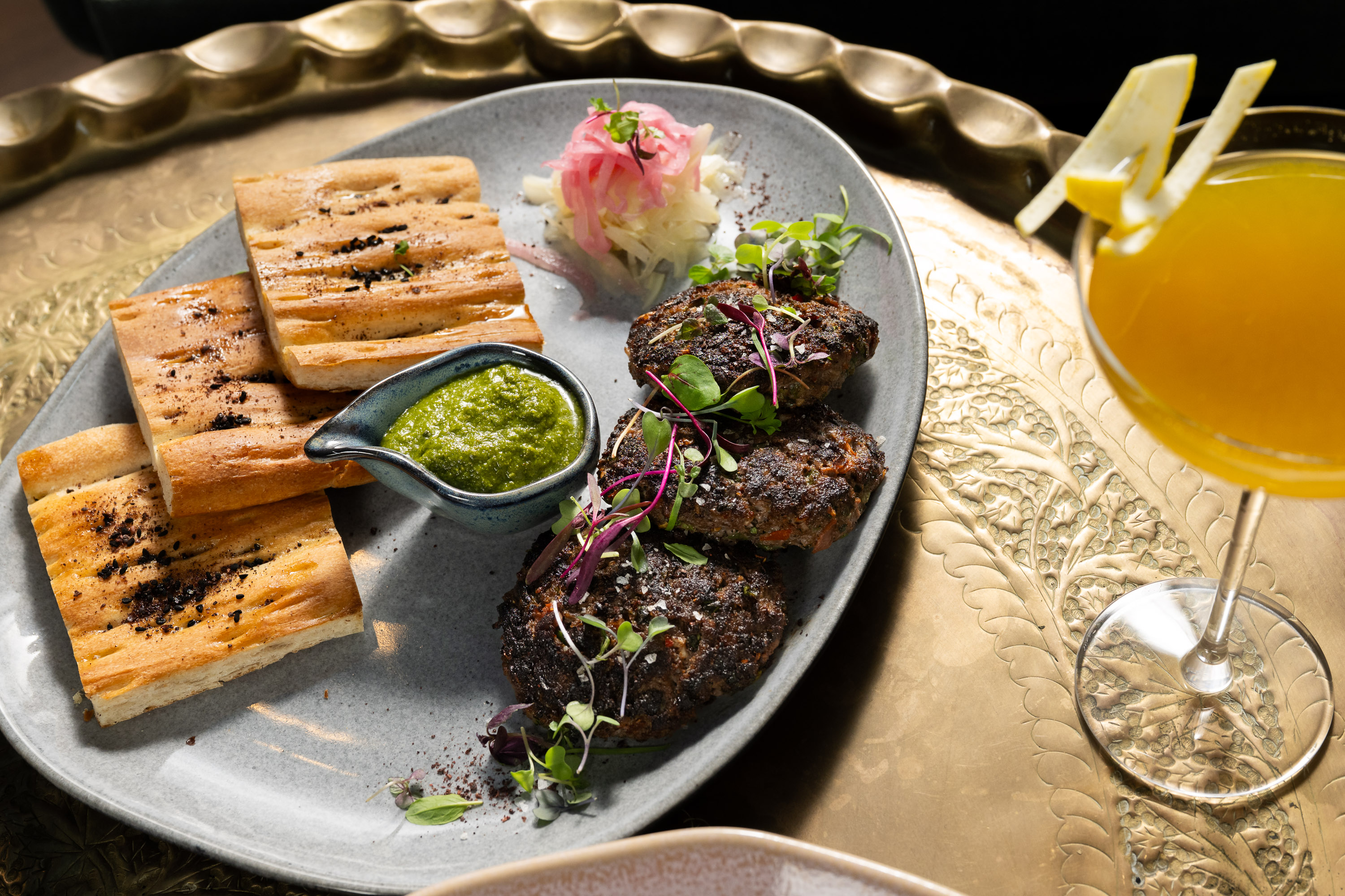 A dish with grilled bread slices, three pieces of seasoned meat, green sauce, and pickled onions sits on a tray, next to a cocktail with a lemon twist.
