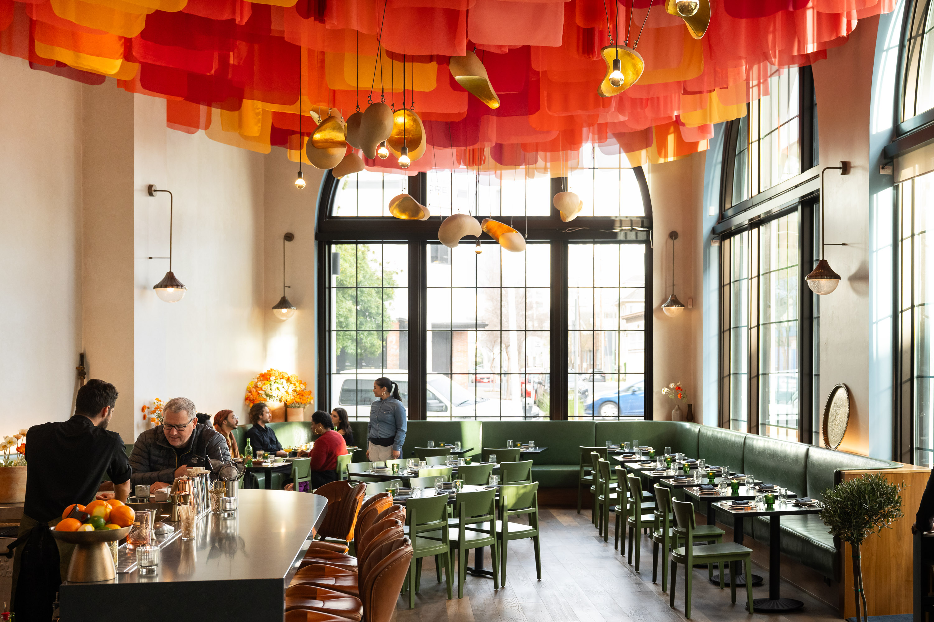 A bright restaurant has colorful fabric hanging from the ceiling, large windows, and green chairs around tables. People are seated and chatting, and there's a bar area.