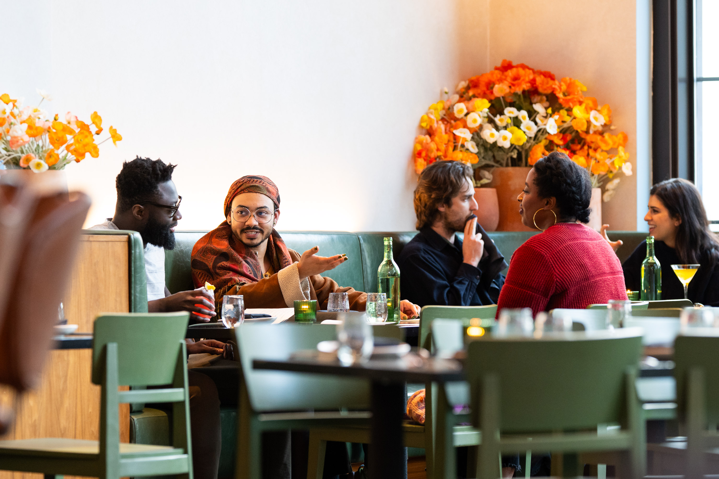 A group of six people are engaged in conversation around a restaurant table. The setting is cozy with vibrant orange and white flowers in the background.