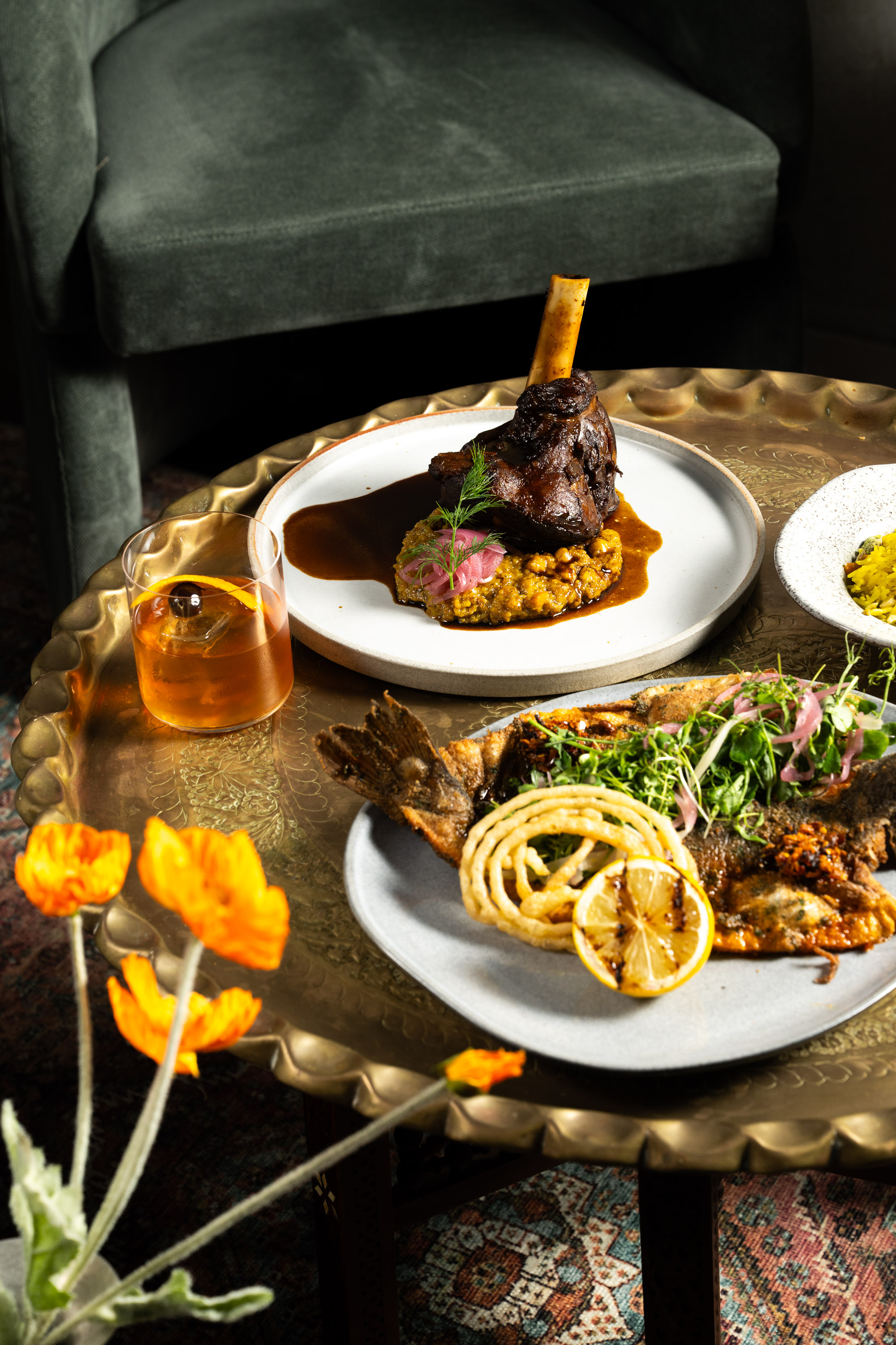 A brass table holds a gourmet meal with roasted lamb on couscous, whole fish garnished with lemon and greens, and an orange cocktail, framed by orange flowers.