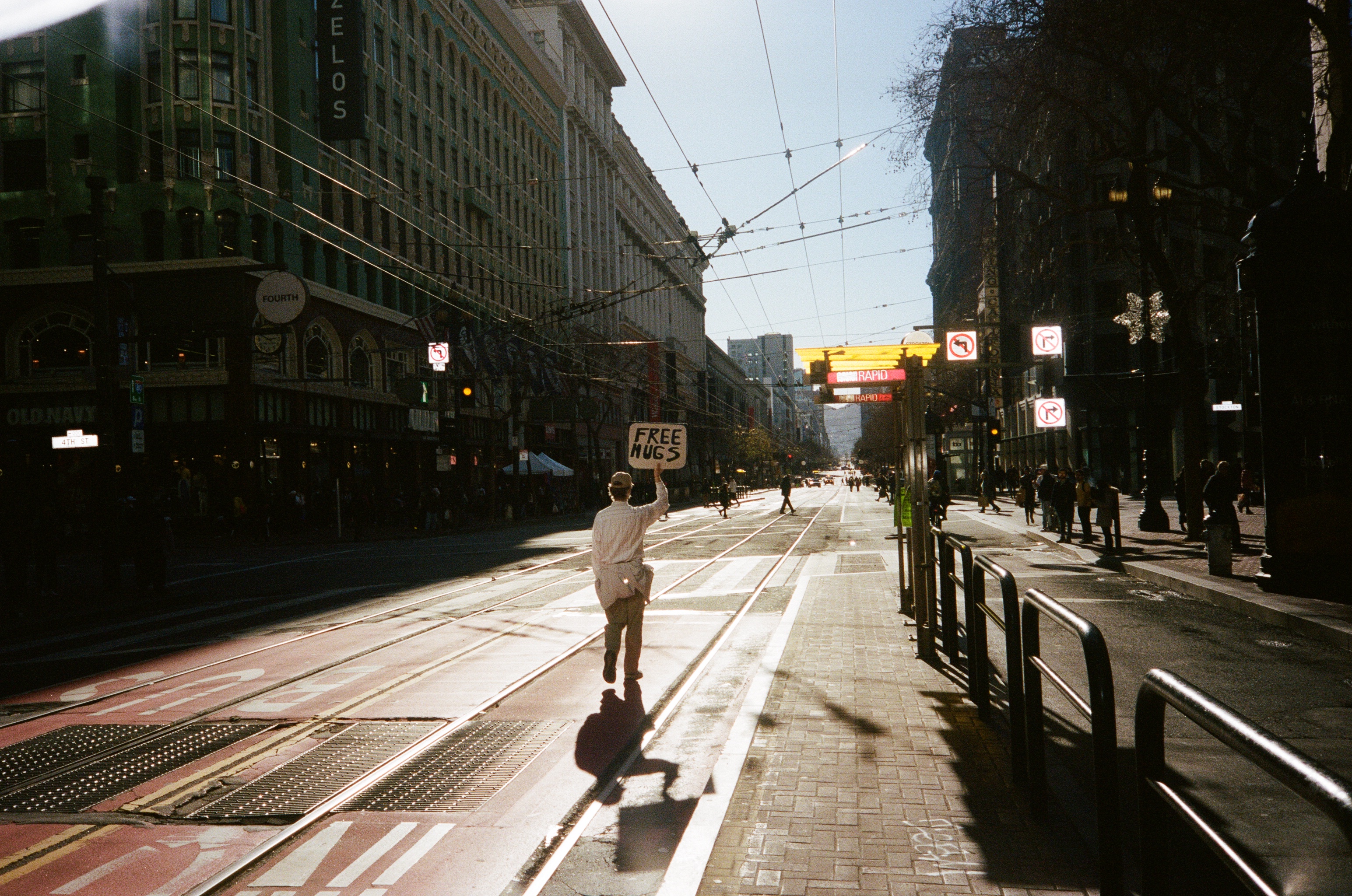 A person walks down a city street holding a &quot;Free Hugs&quot; sign. The street is lined with buildings and illuminated by bright sunlight.