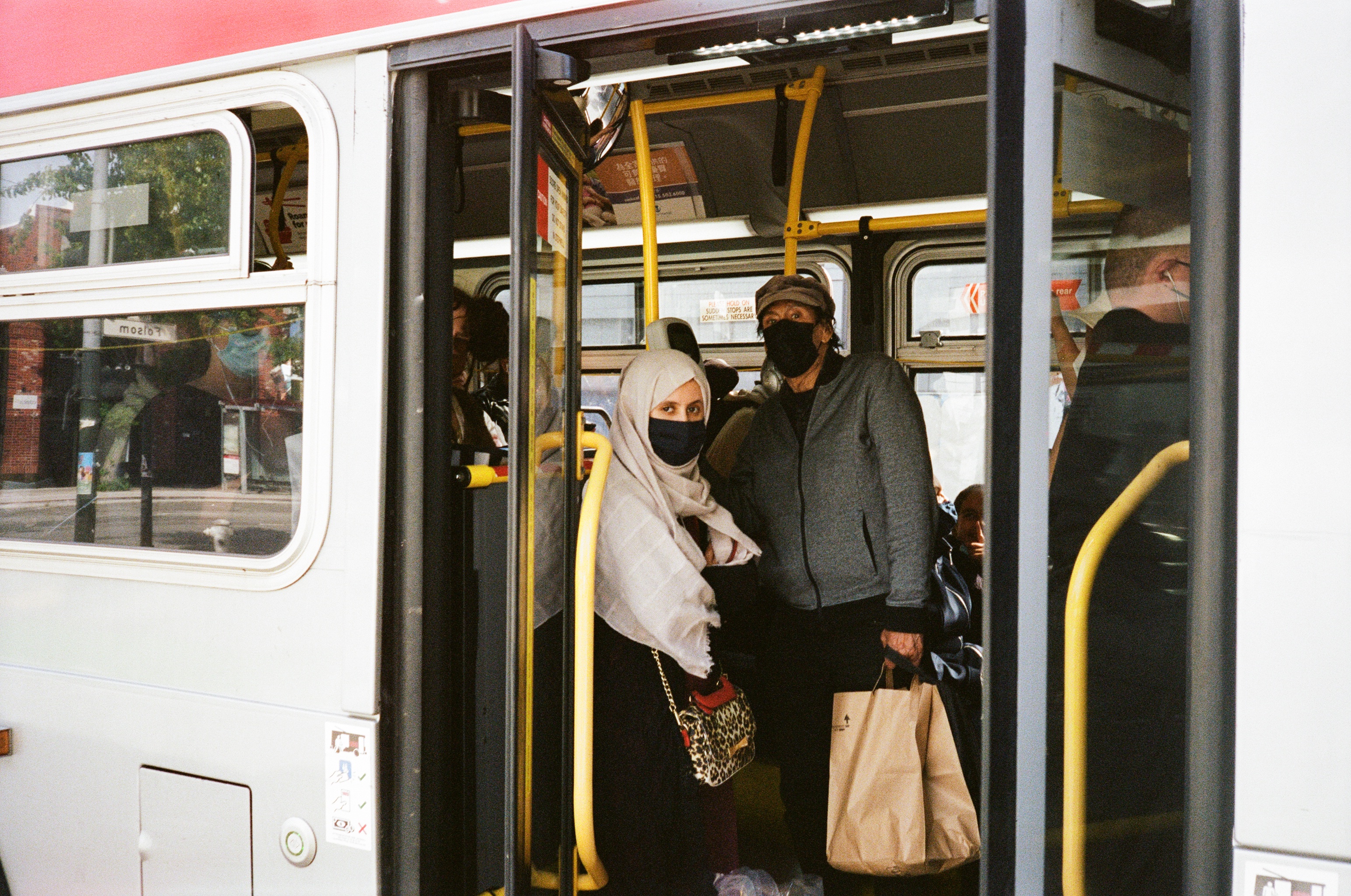 Two people wearing masks are standing inside a bus by the door. One person is carrying a shopping bag, and the other has a patterned purse. The bus interior is visible.