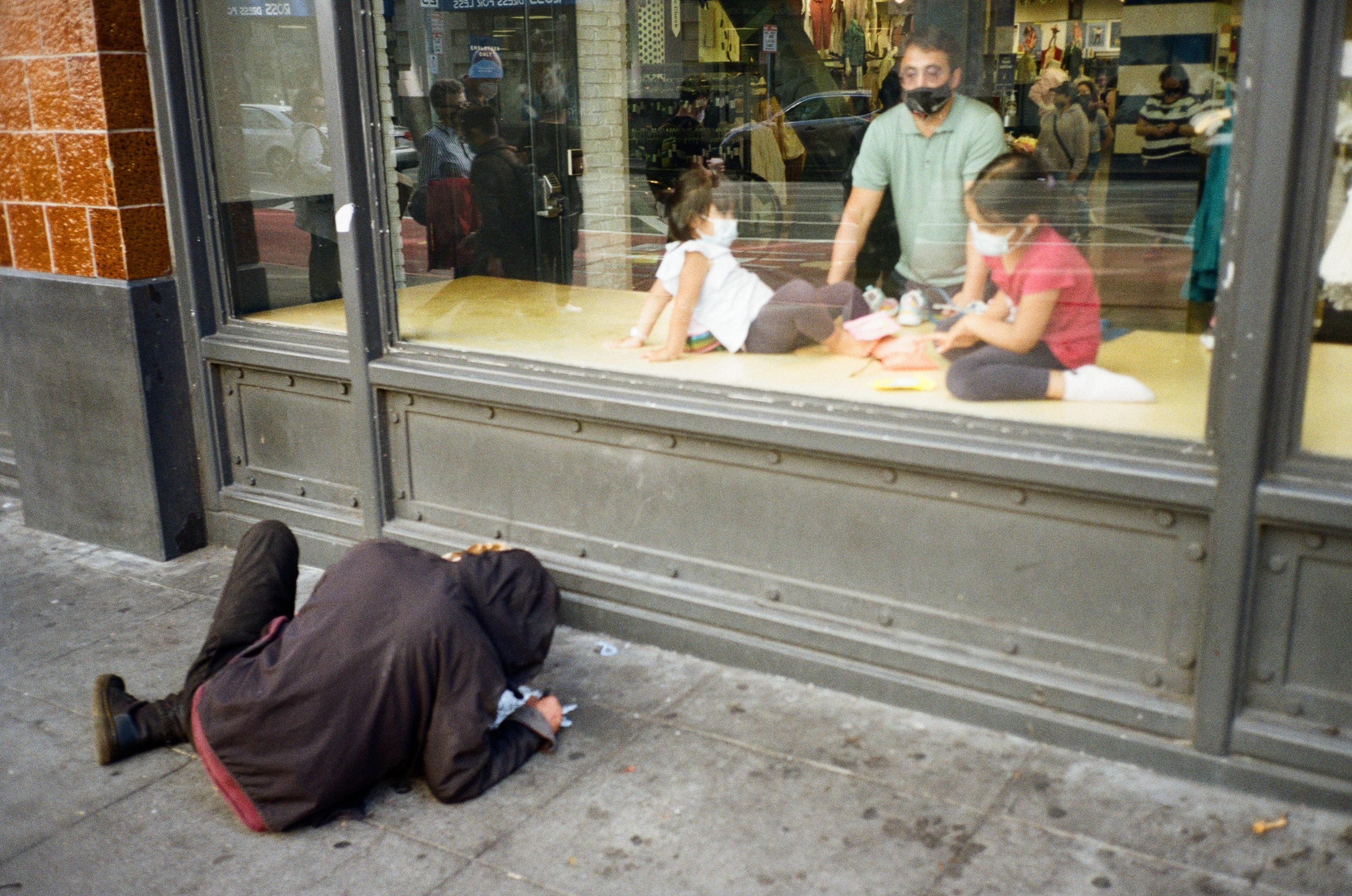 A person in a hooded jacket is lying on a sidewalk beside a storefront window. Inside, a man and two children, all wearing masks, are visible through the glass.