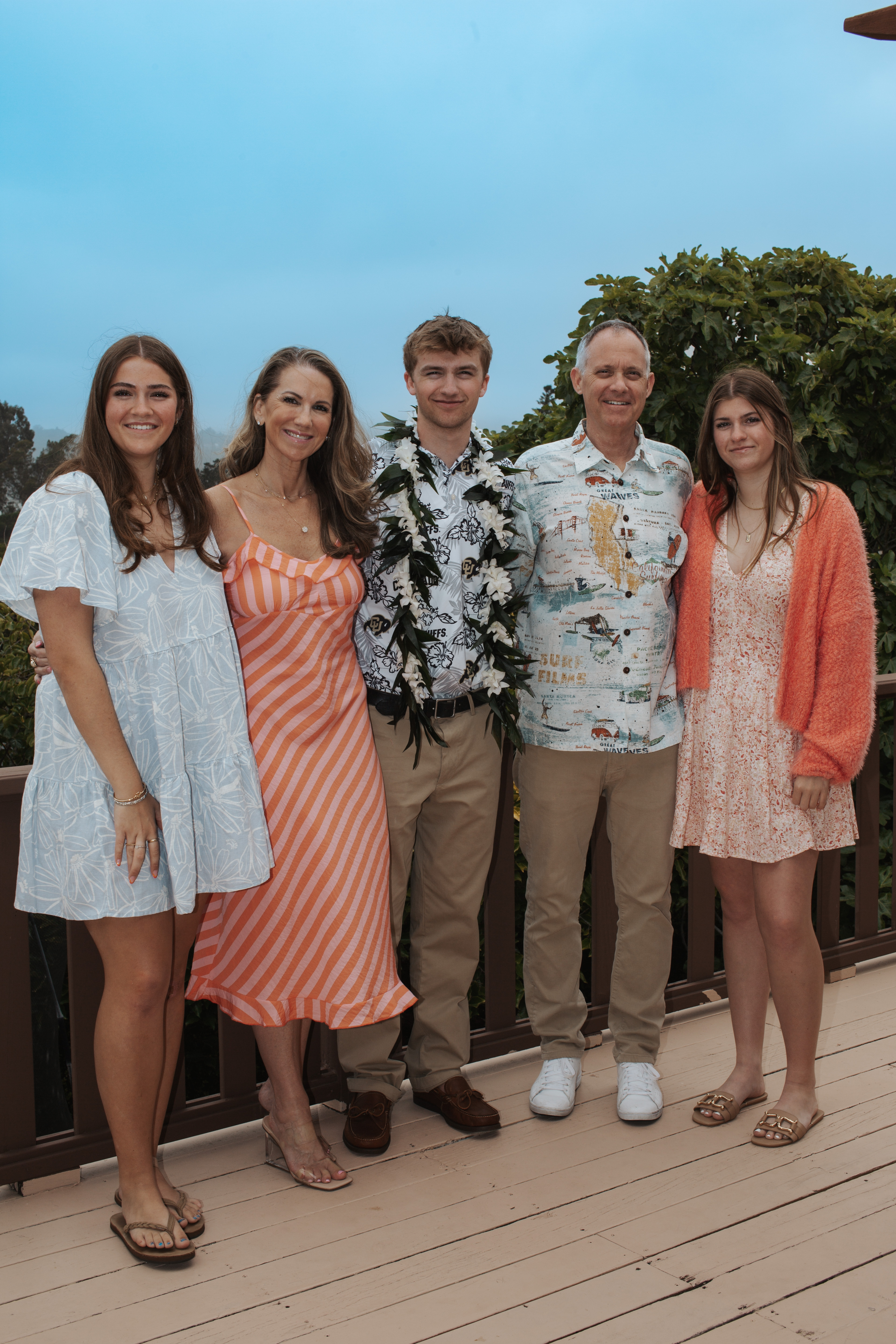 A family of five stands closely on a deck, smiling. They're dressed in summer clothes with a mix of dresses, button-down shirts, and leis. A lush background is visible.