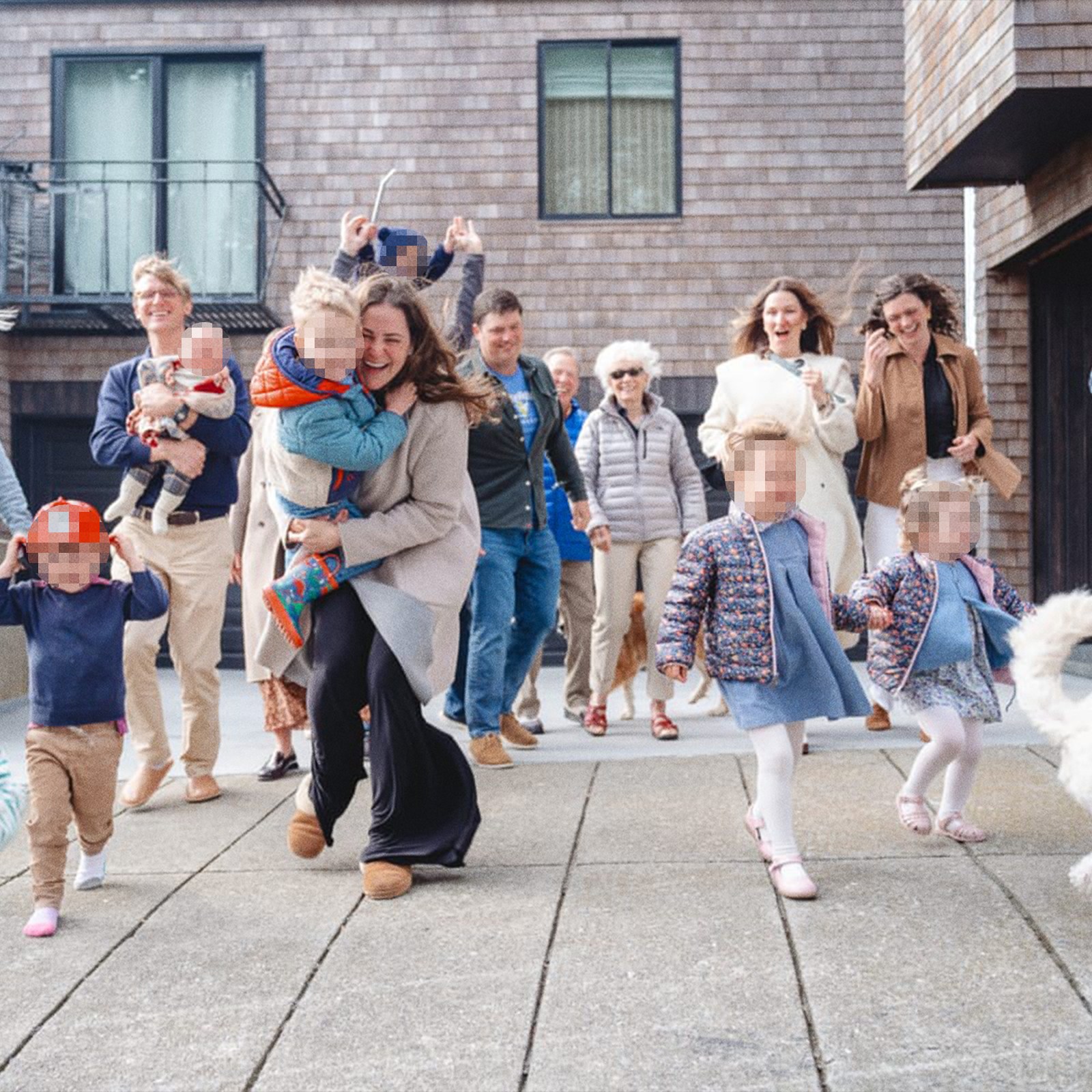 A joyful group of adults and children, some with pixelated faces, are outside a house, happily running and interacting, with a dog in the foreground.