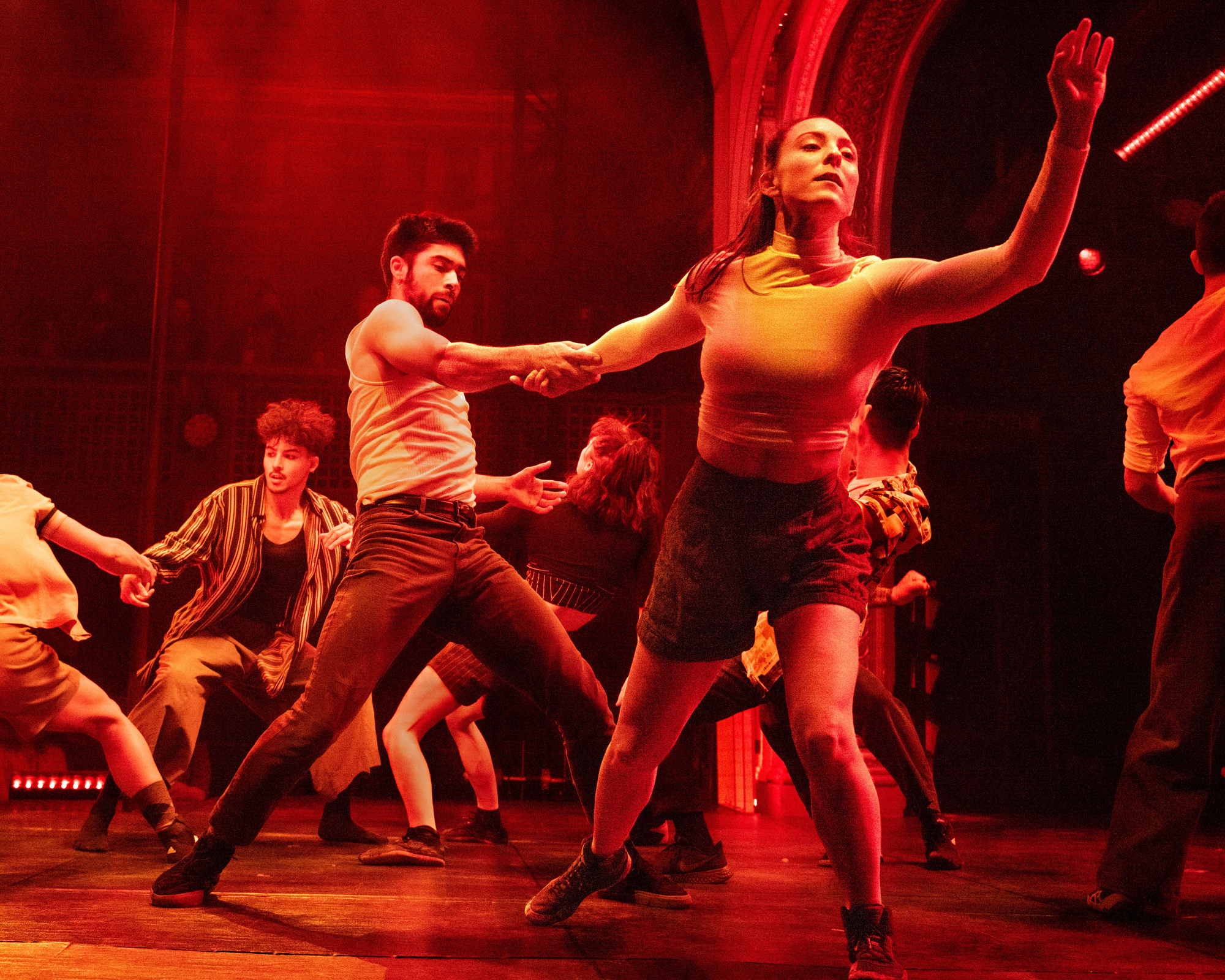 A group of dancers perform energetically on stage with warm, dramatic lighting. A male dancer in the foreground holds hands with a female dancer, both in dynamic poses.