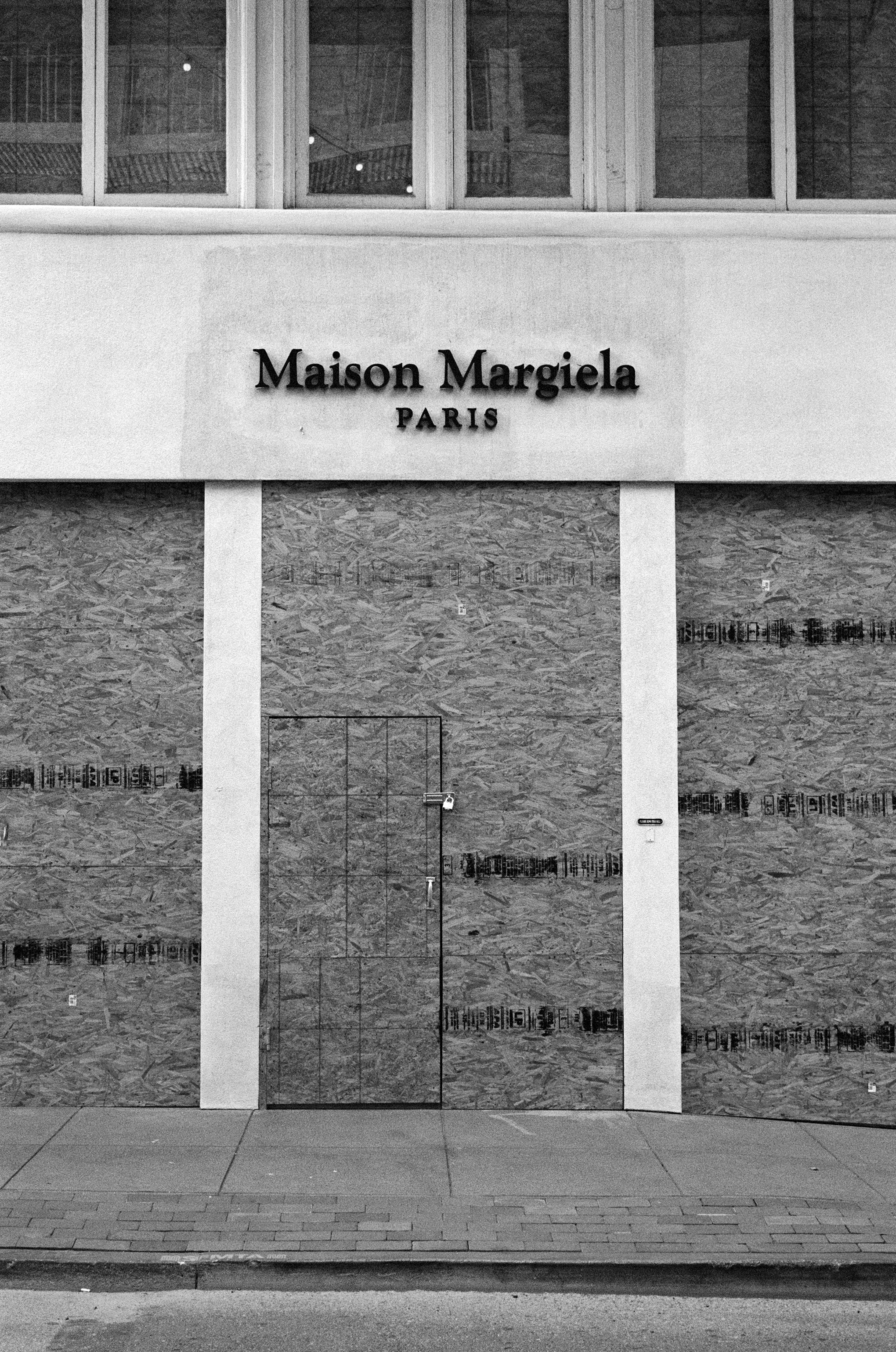 The image shows a closed storefront, boarded with wood panels. Above, &quot;Maison Margiela Paris&quot; is written in black letters on a light wall.