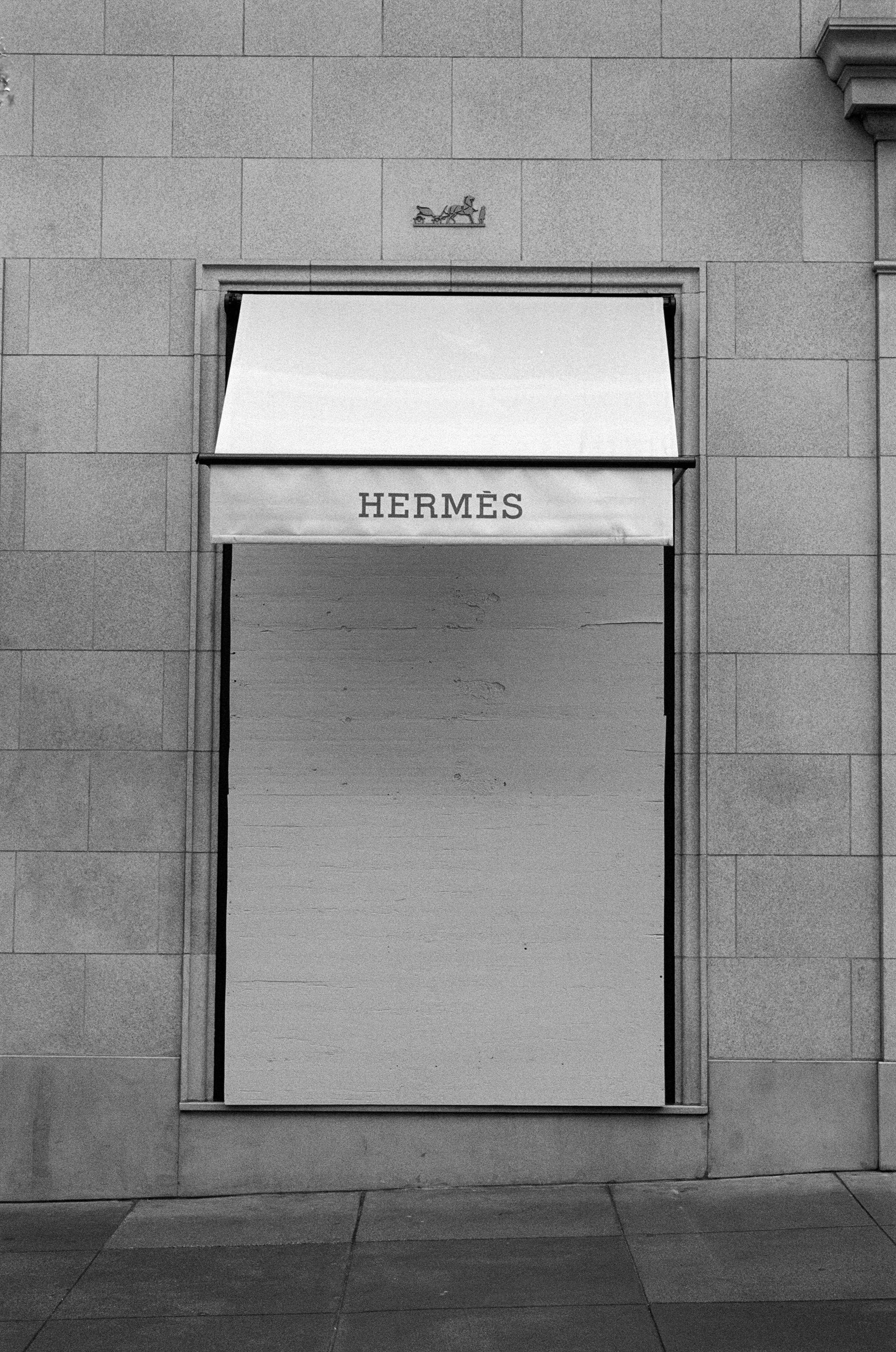 A boarded-up storefront with a &quot;Hermès&quot; awning. The wall is made of large stone tiles, and there's a simple design above the awning.