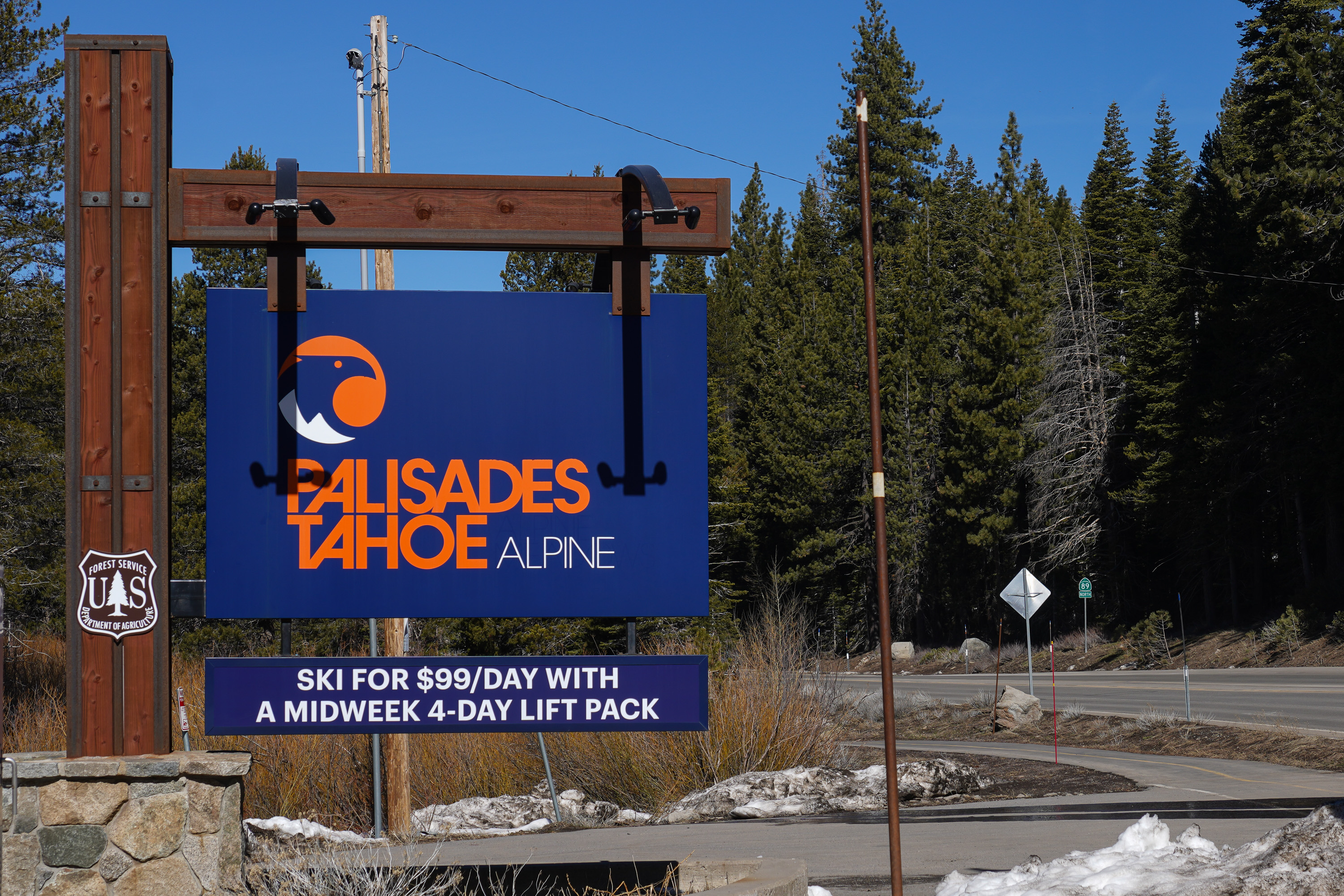 A sign reads &quot;Palisades Tahoe Alpine&quot; with an offer to ski for $99/day with a midweek 4-day lift pack. It's set in a forested area by a road.