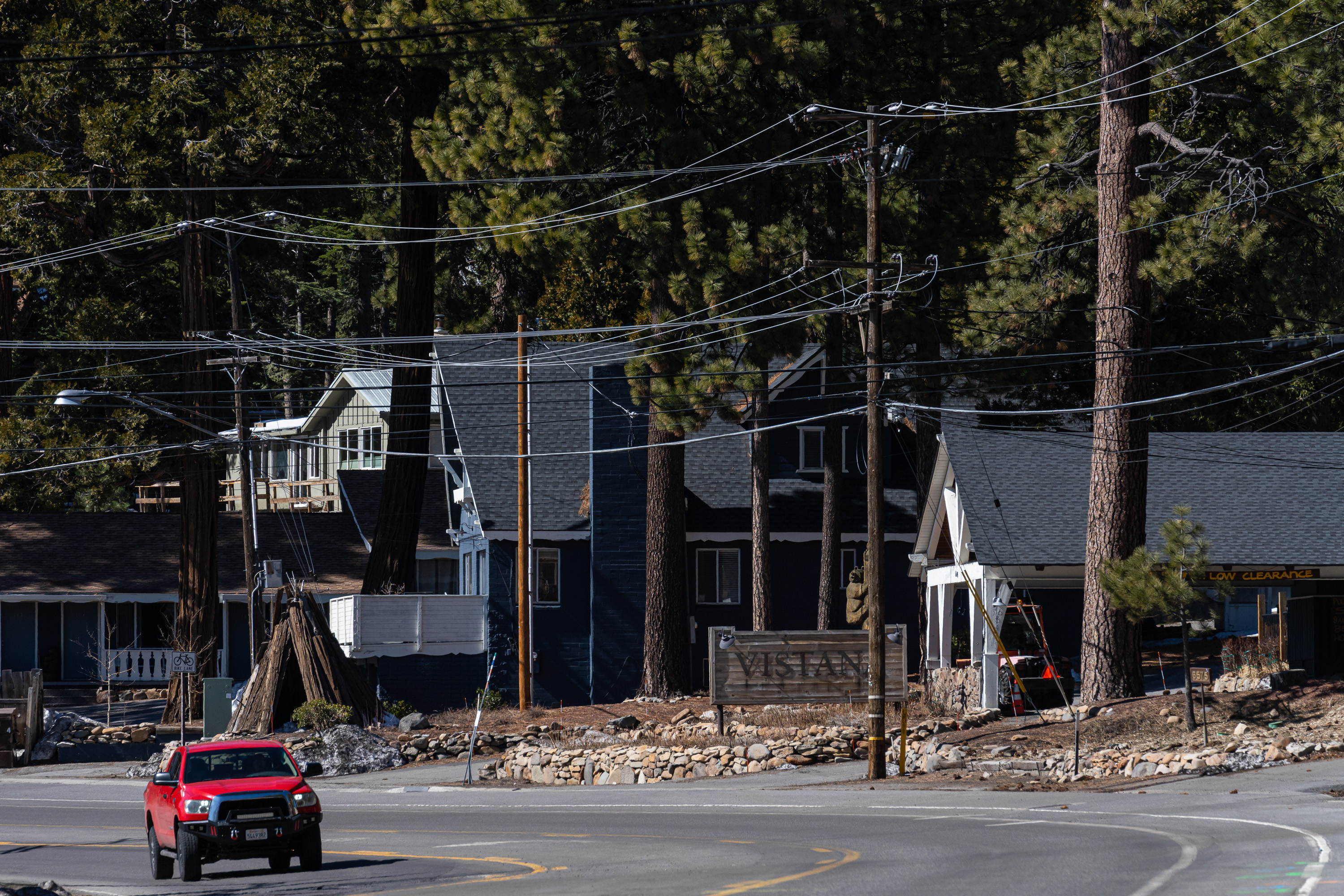 A red truck drives past a sign reading &quot;VISTAN&quot; in a wooded area with tall trees. There are houses surrounded by power lines and pine trees.