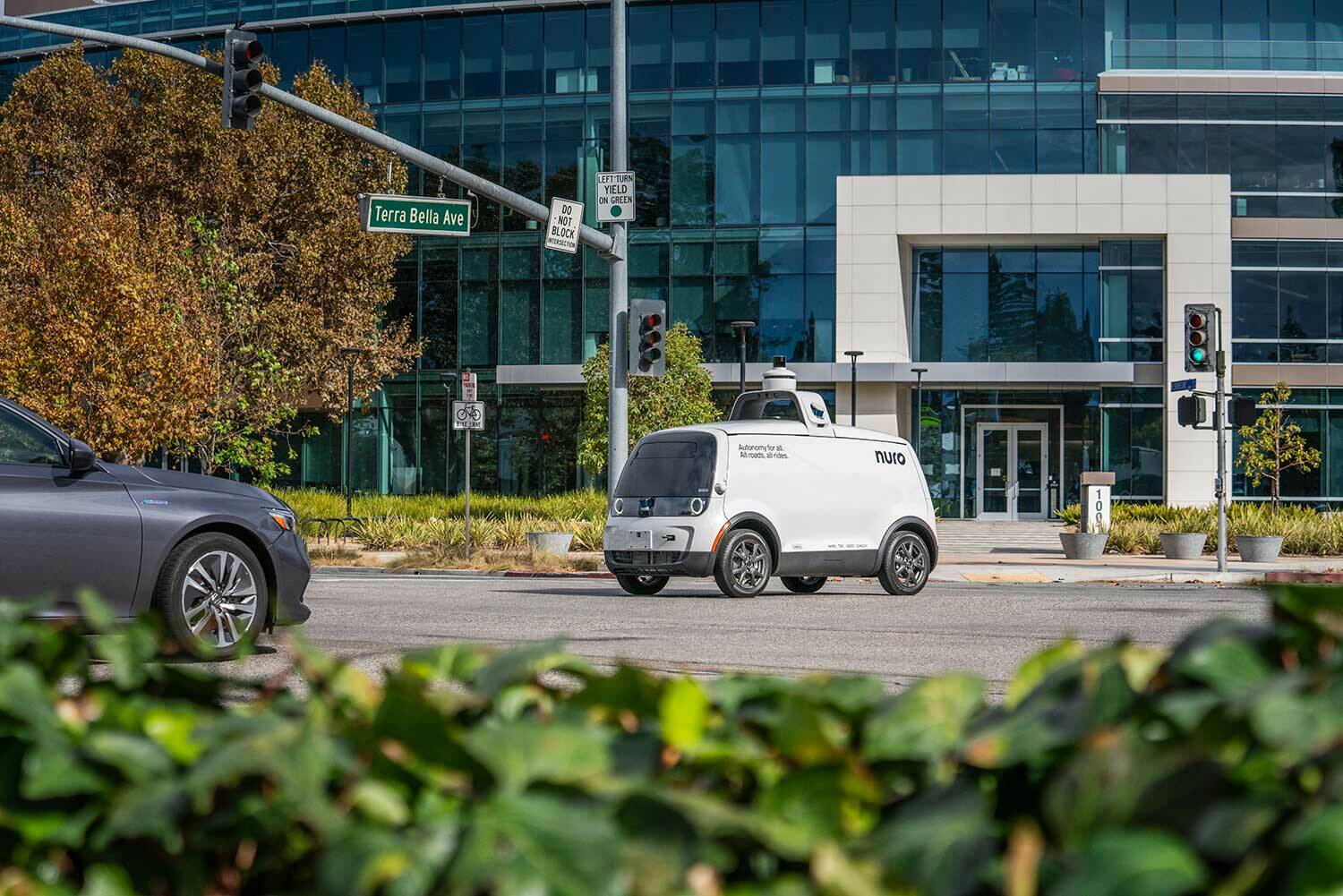 A small autonomous delivery vehicle is driving on a street near a modern glass building. Traffic lights and a street sign for &quot;Terra Bella Ave&quot; are visible.