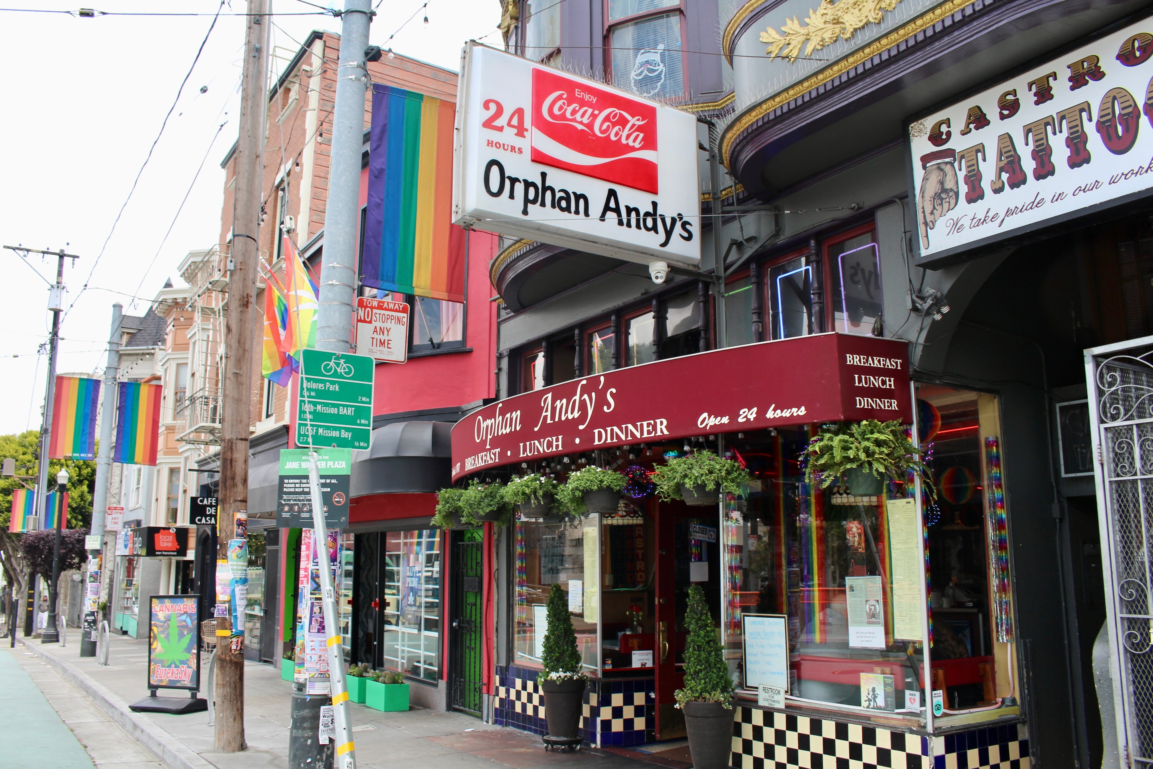 The image shows a vibrant street scene with rainbow flags. "Orphan Andy's" and "Castro Tattoo" signs are visible. The area is lively and colorful.