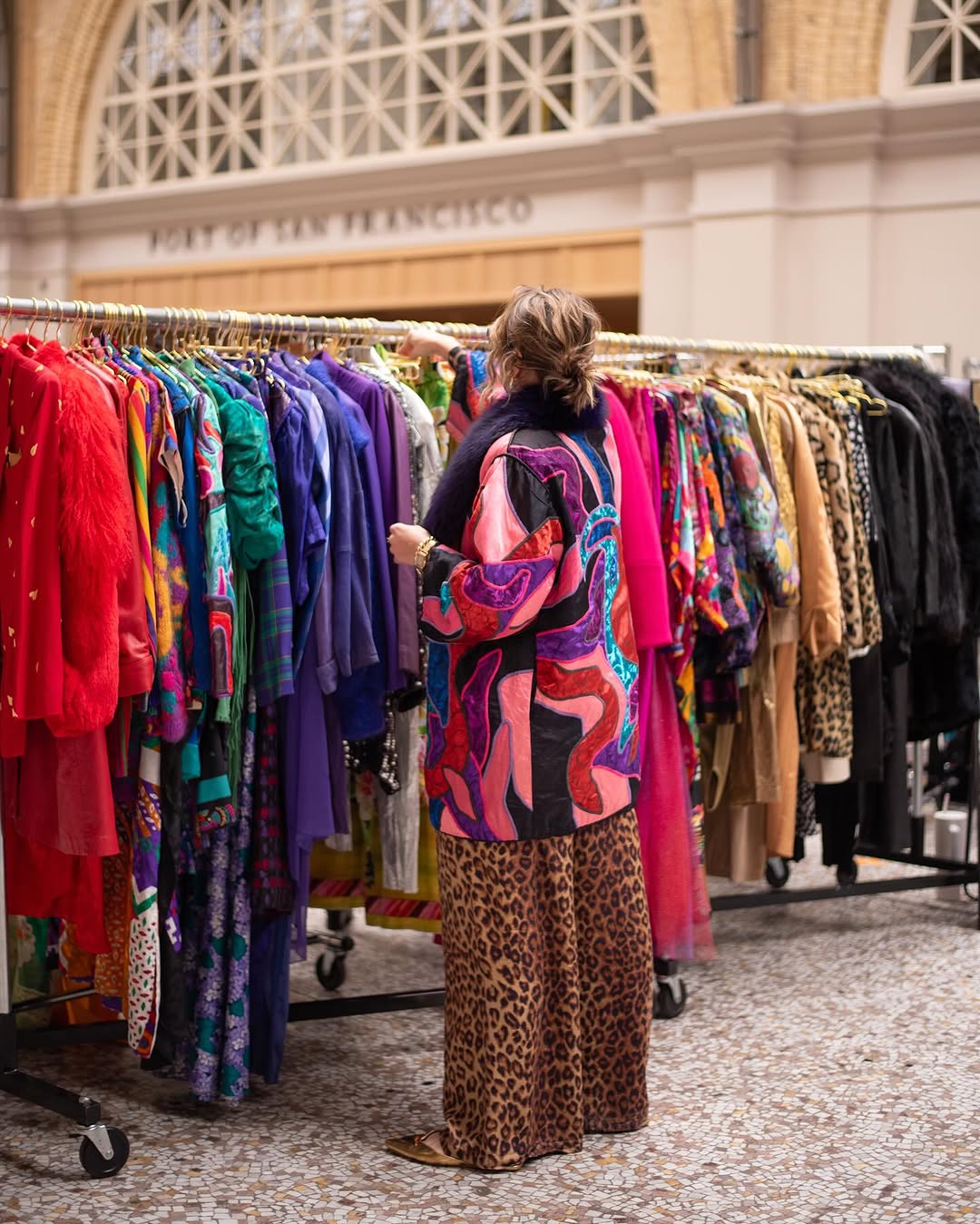 A person wearing a colorful, patterned outfit browses through racks of vibrant, eclectic clothing displayed in front of an arched architectural backdrop.