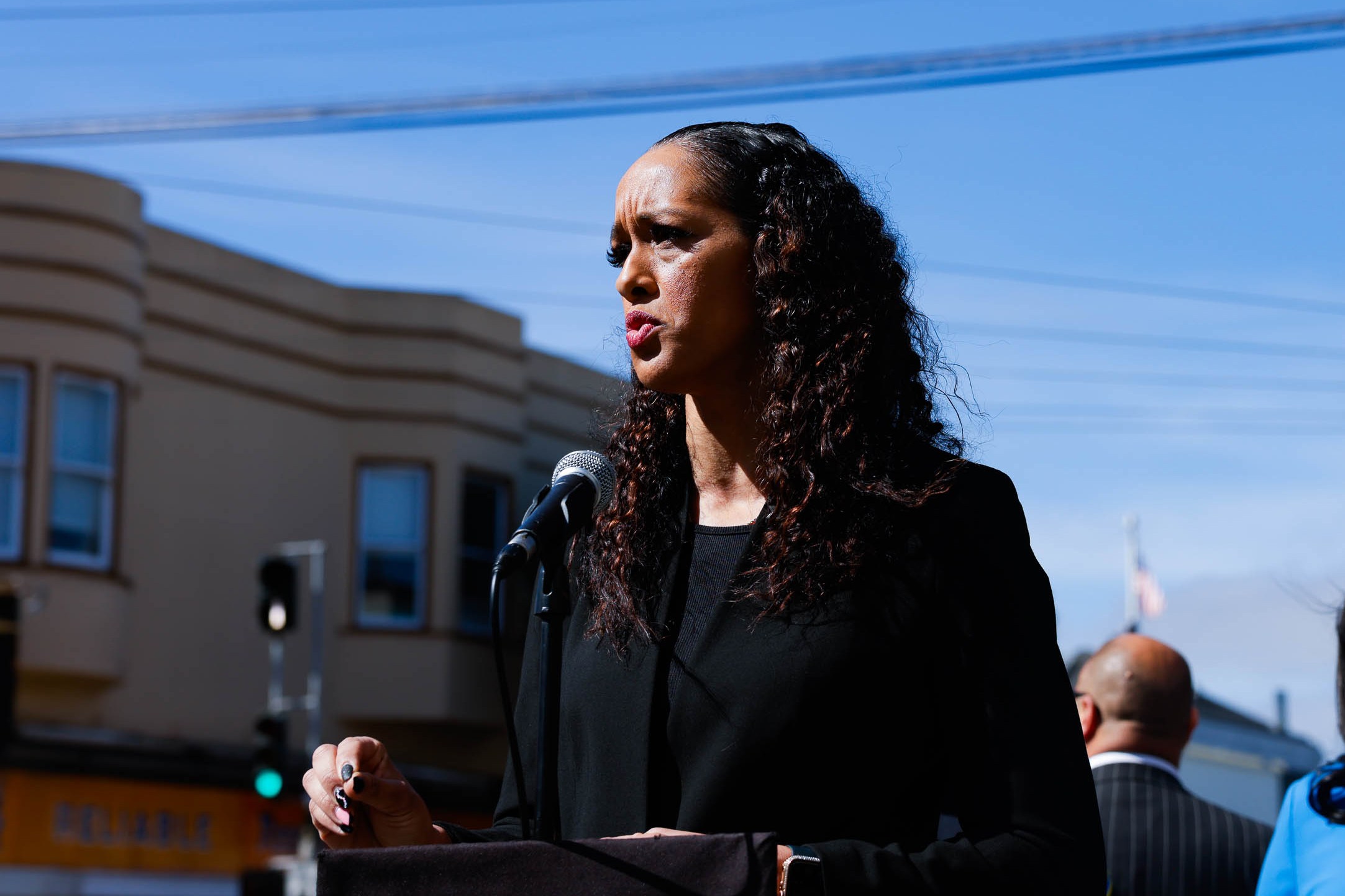 A person with long curly hair speaks into a microphone, standing outdoors on a sunny day. They are in front of a building with large windows.