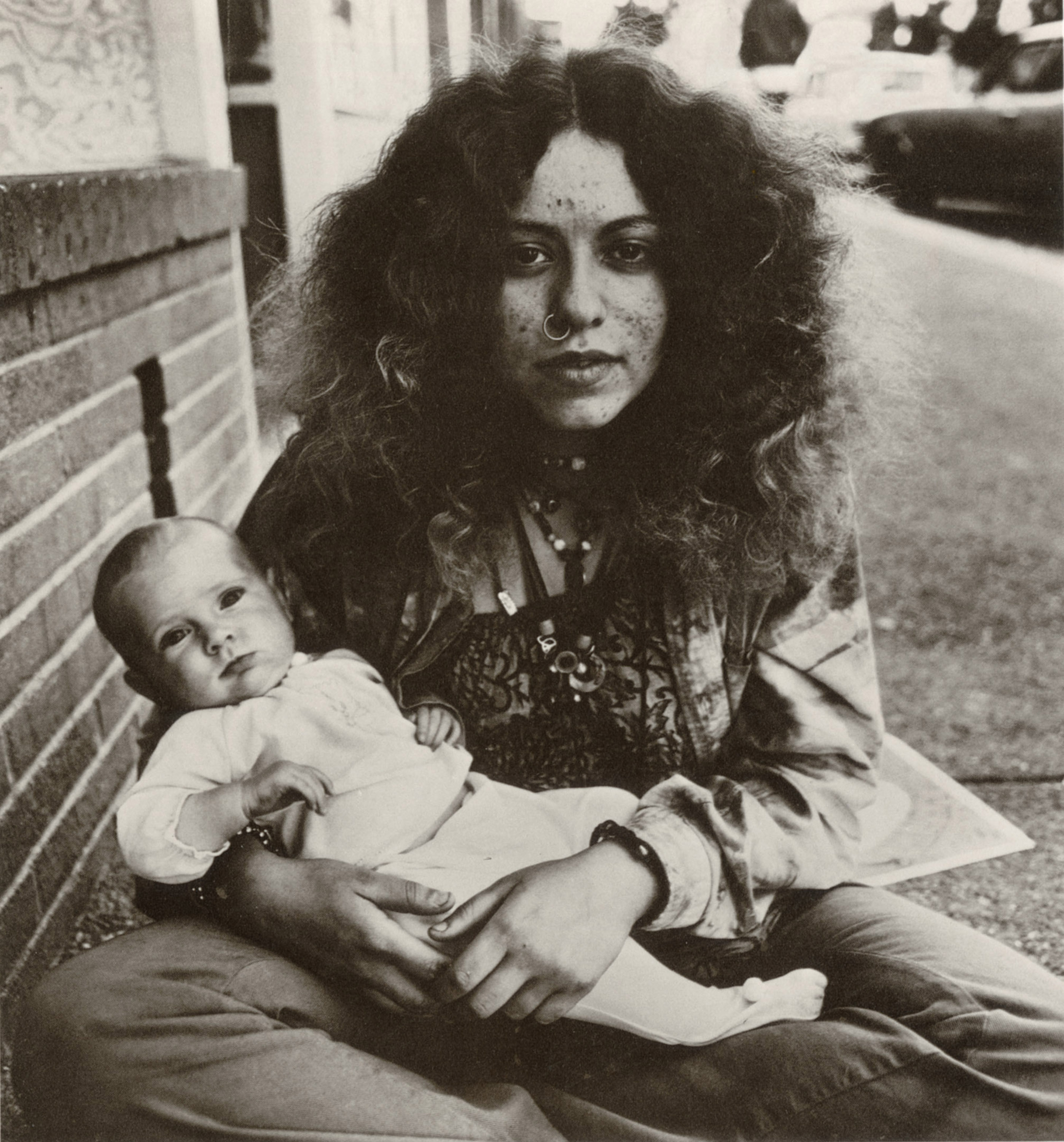 A woman with curly hair, wearing a nose ring and necklaces, sits on a sidewalk holding a baby. Both look directly at the camera. A brick wall is behind them.