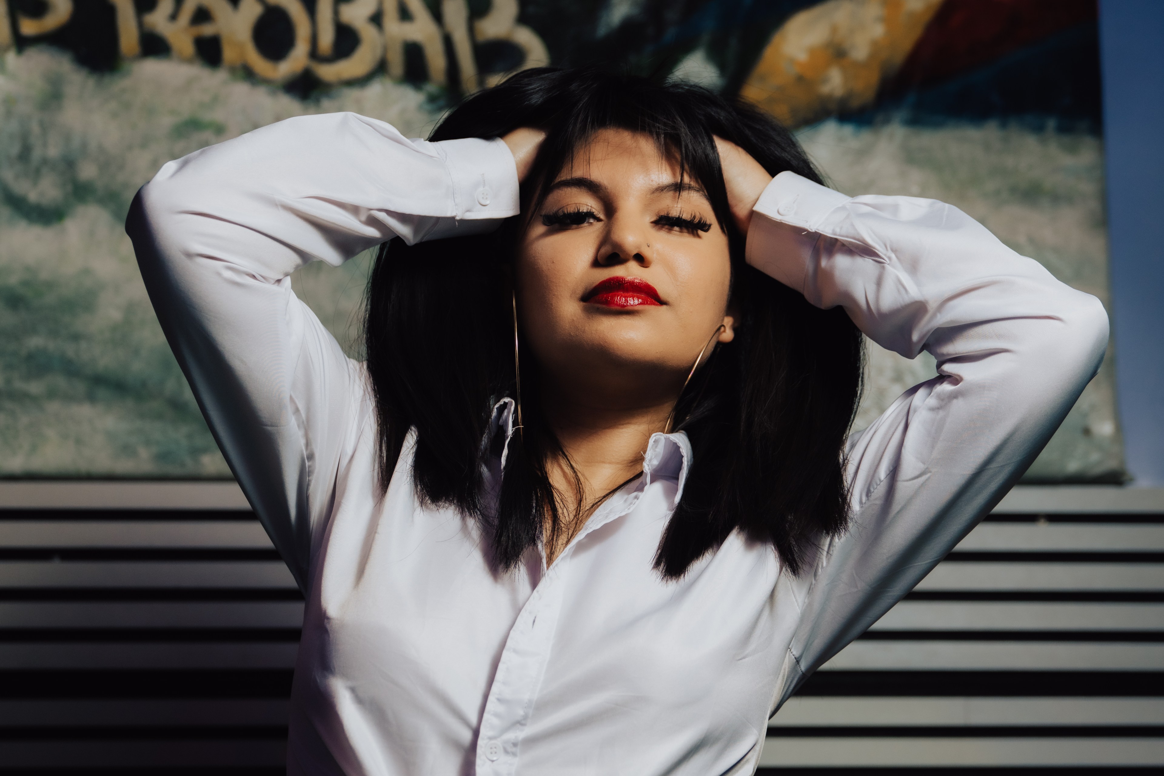 A woman with dark hair poses confidently with hands on her head. She's wearing a tied white shirt and jeans. A colorful, abstract background is behind her.