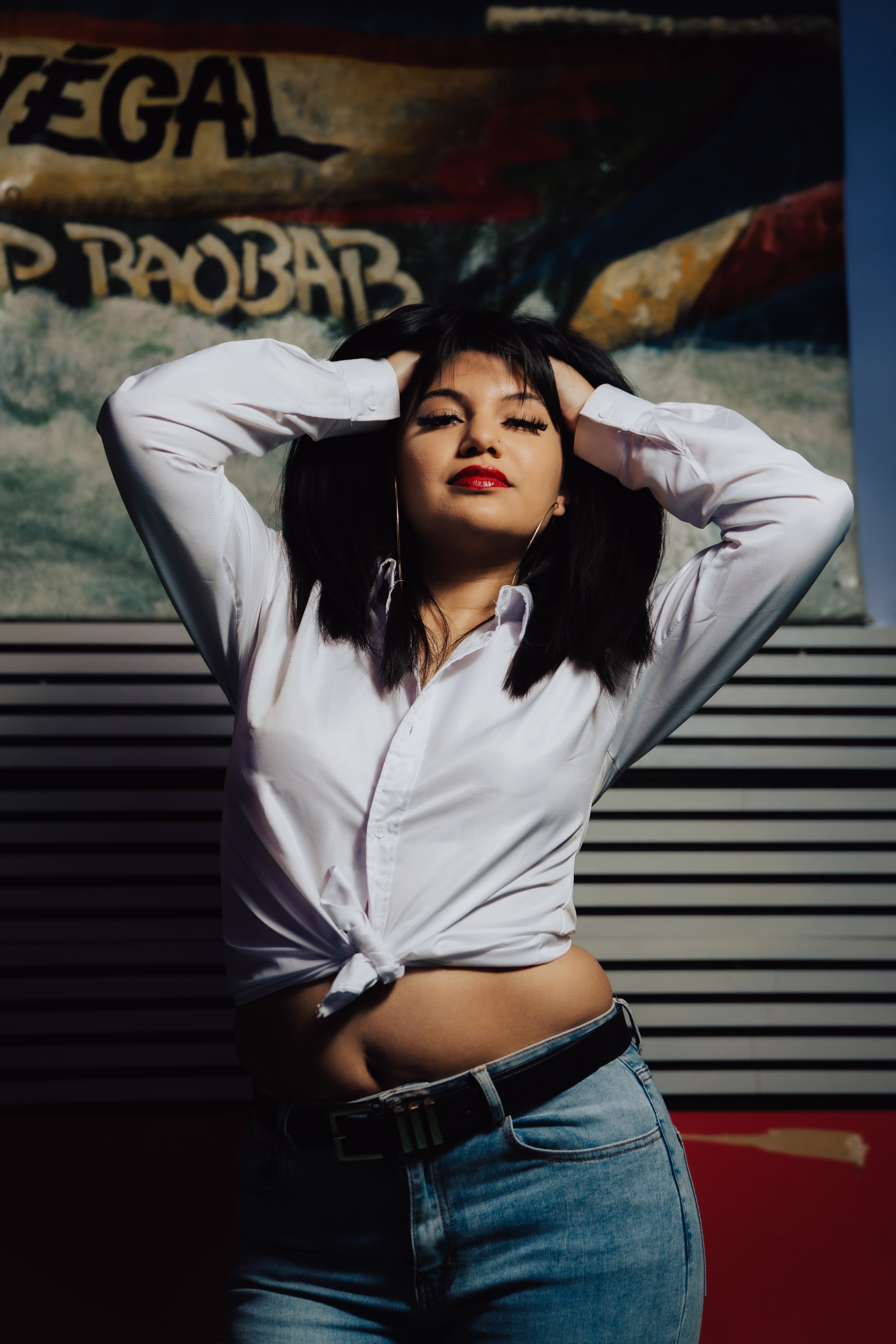 A woman with dark hair poses confidently with hands on her head. She's wearing a tied white shirt and jeans. A colorful, abstract background is behind her.