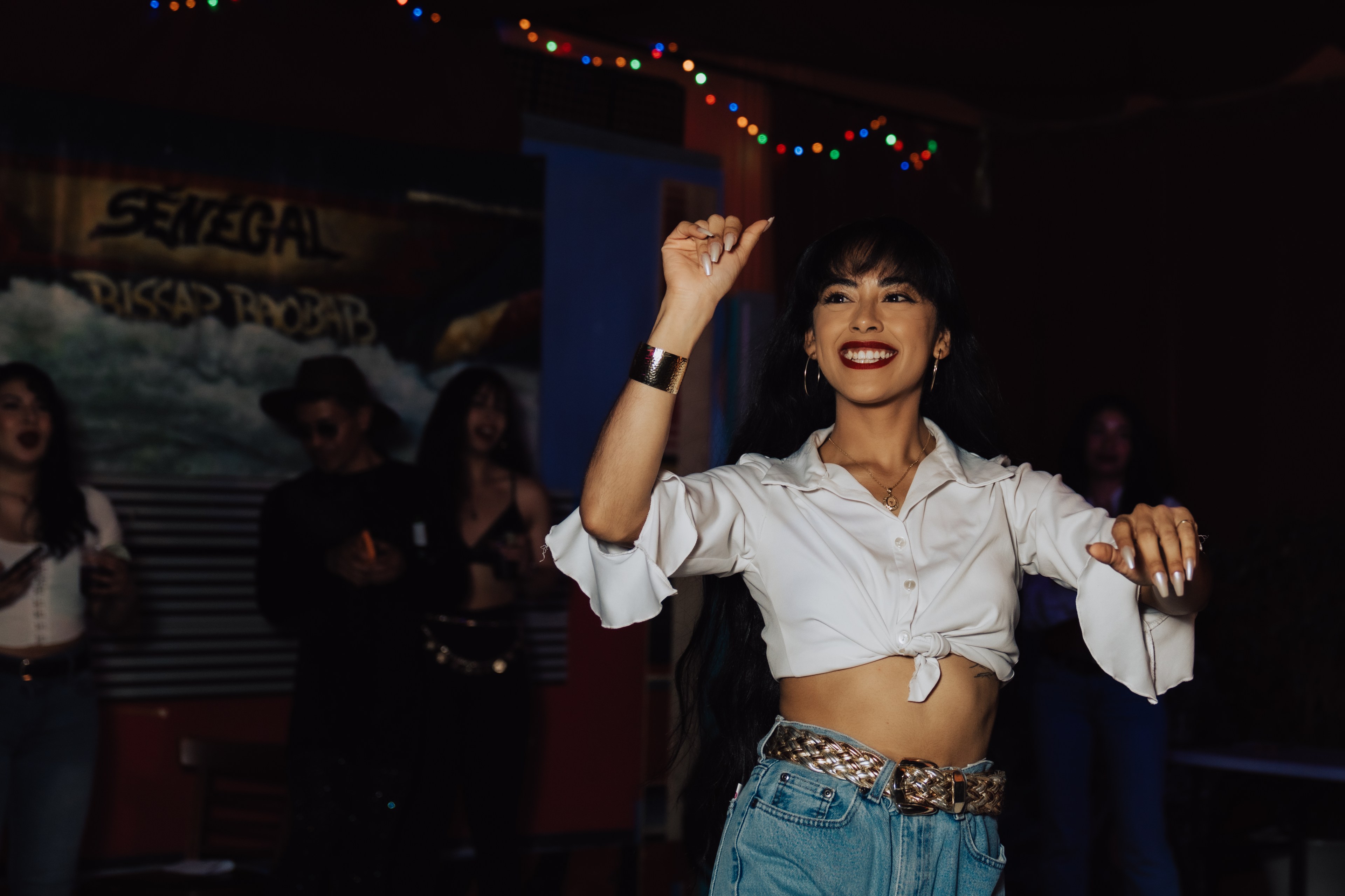 A woman in a white blouse and jeans joyfully dances, surrounded by festively lit, colorful lights. She's smiling brightly, with others blurred in the background.