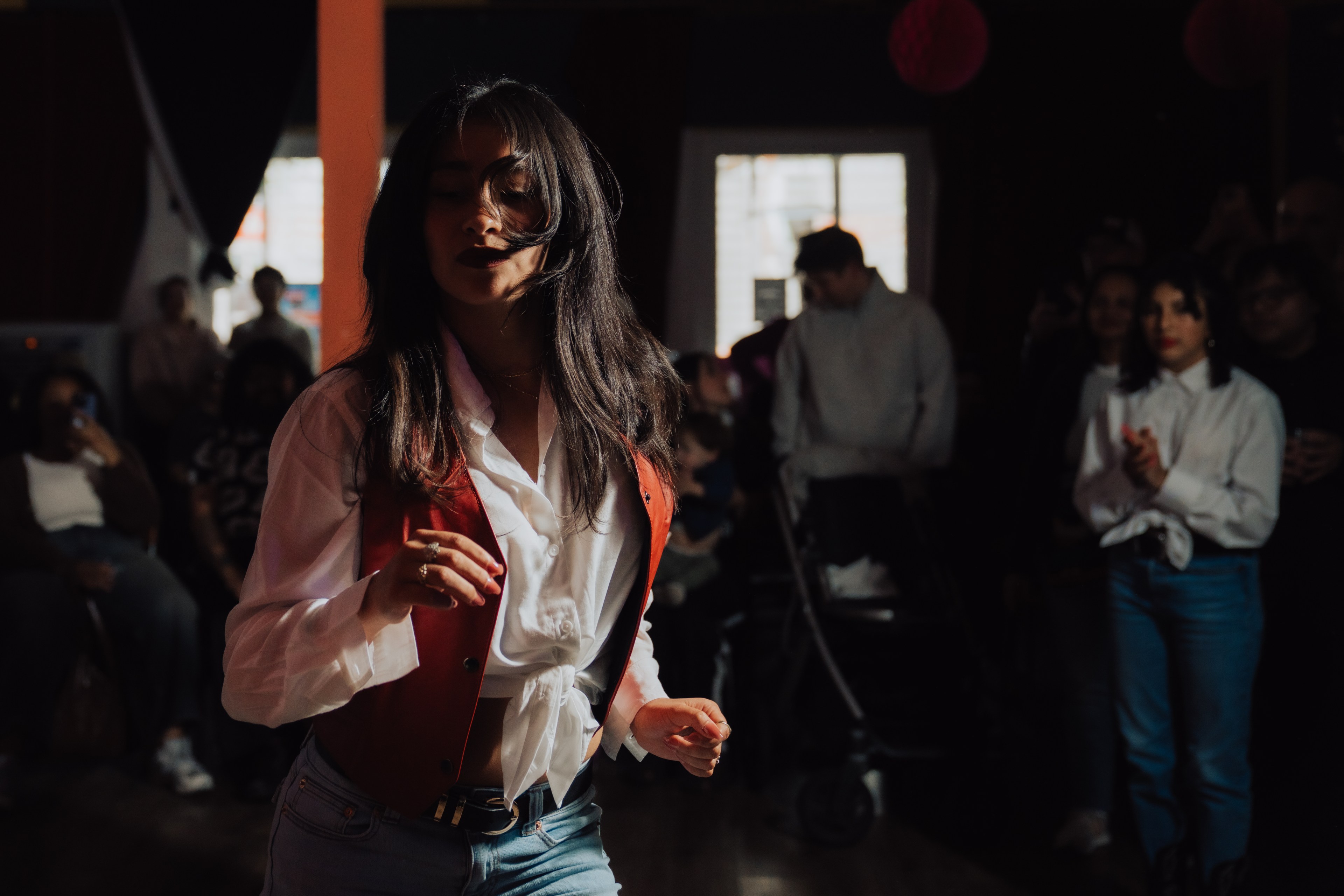 In a dimly lit room, a person in a white shirt and red vest dances energetically. A crowd watches, and some individuals are seated or standing in the background.