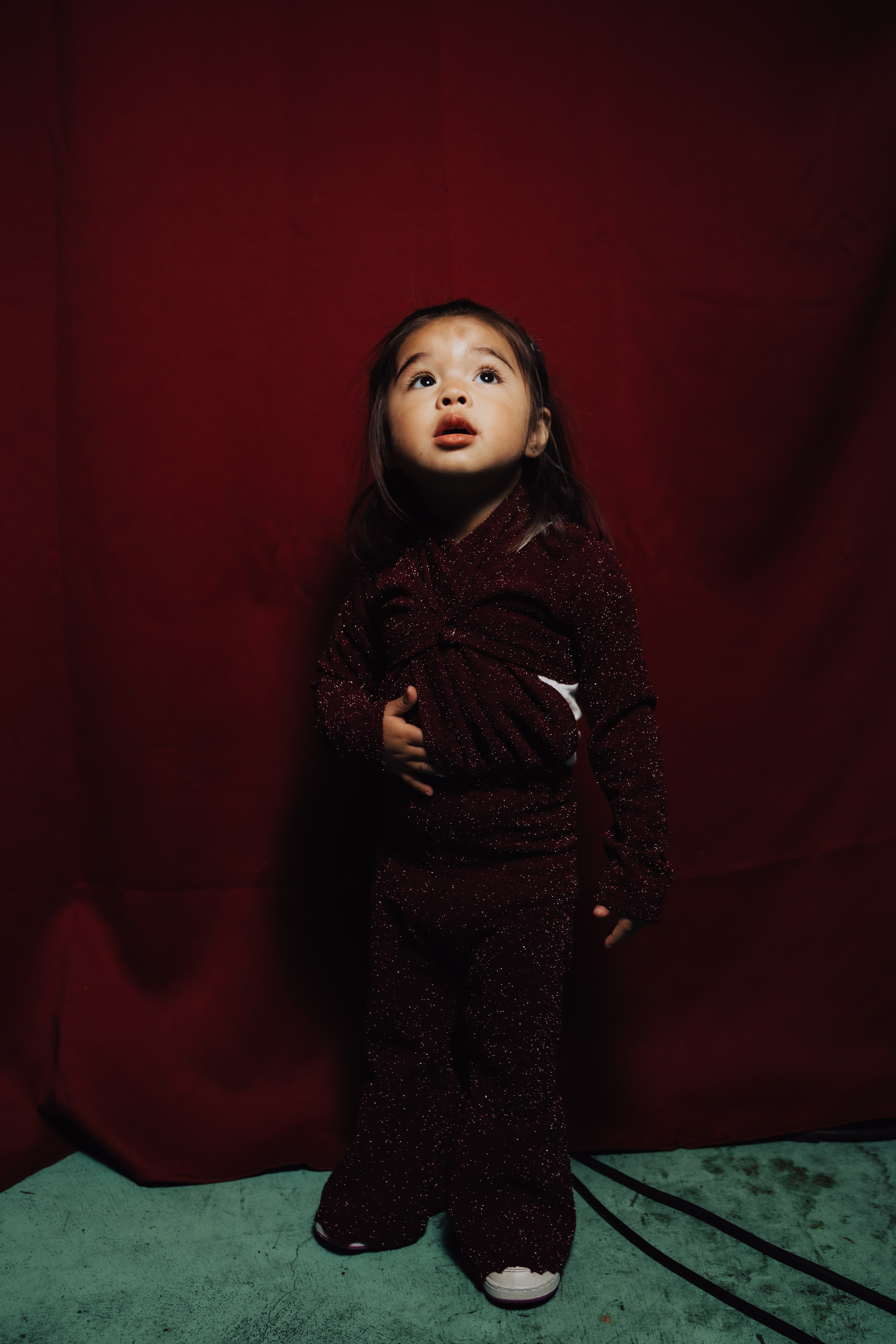 A young child stands against a deep red backdrop, wearing a sparkly, maroon outfit. They gaze upward, with one hand on their chest, on a green surface.