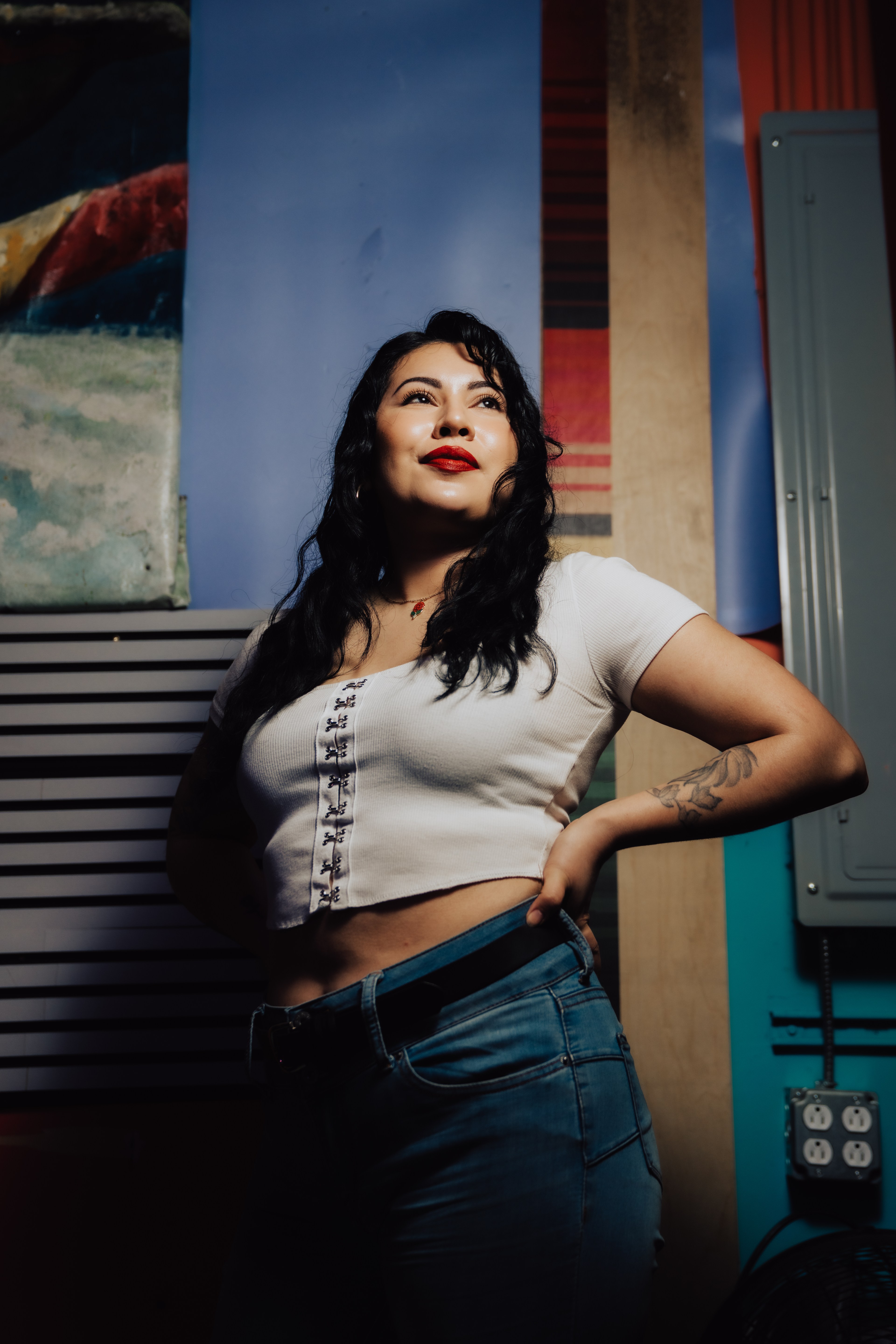 A woman with long dark hair stands confidently, wearing a white crop top and jeans. She's posing with hands on her hips against a colorful, artistic backdrop.