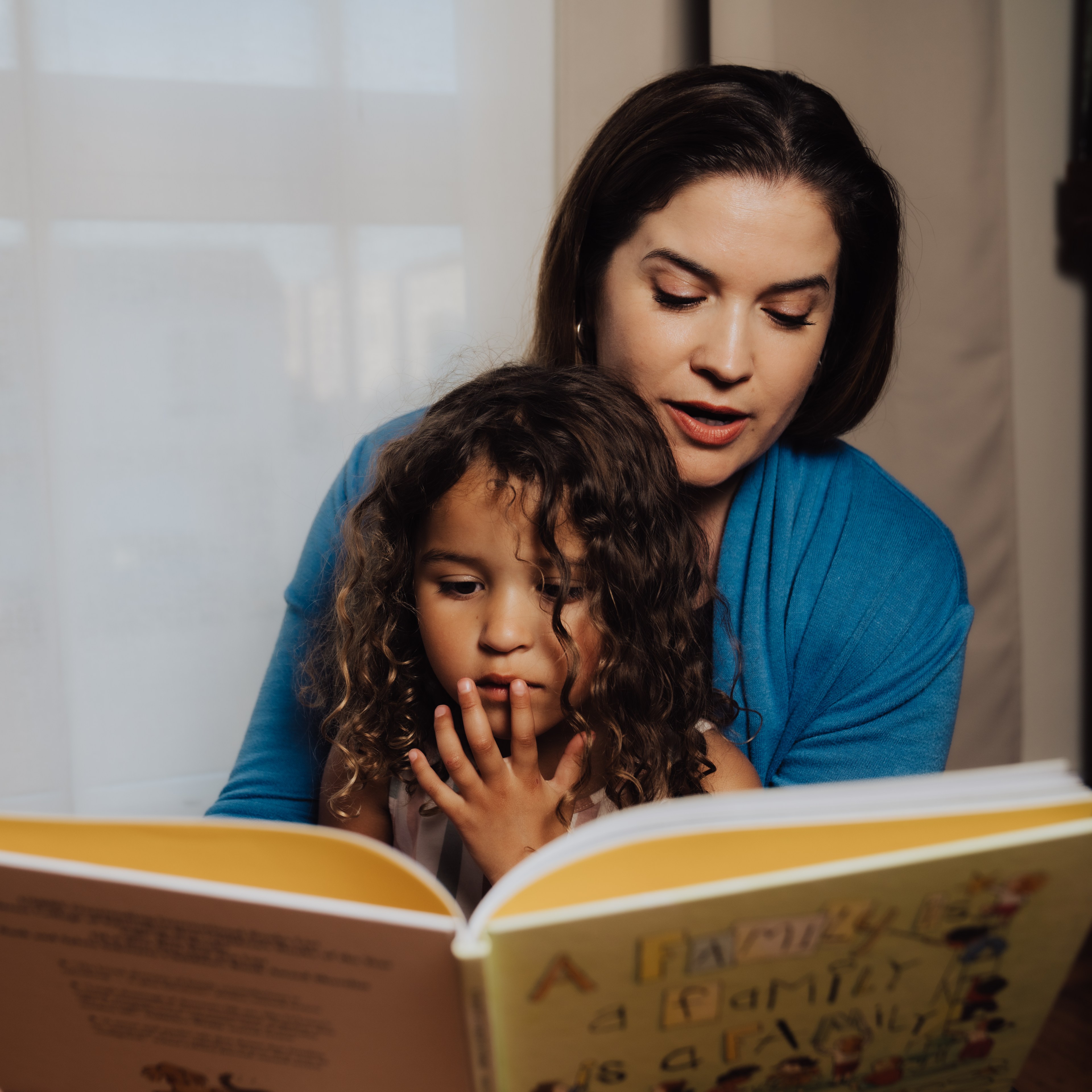 A woman in a blue sweater reads a colorful book to a young girl with curly hair. They sit closely together in a softly lit room.