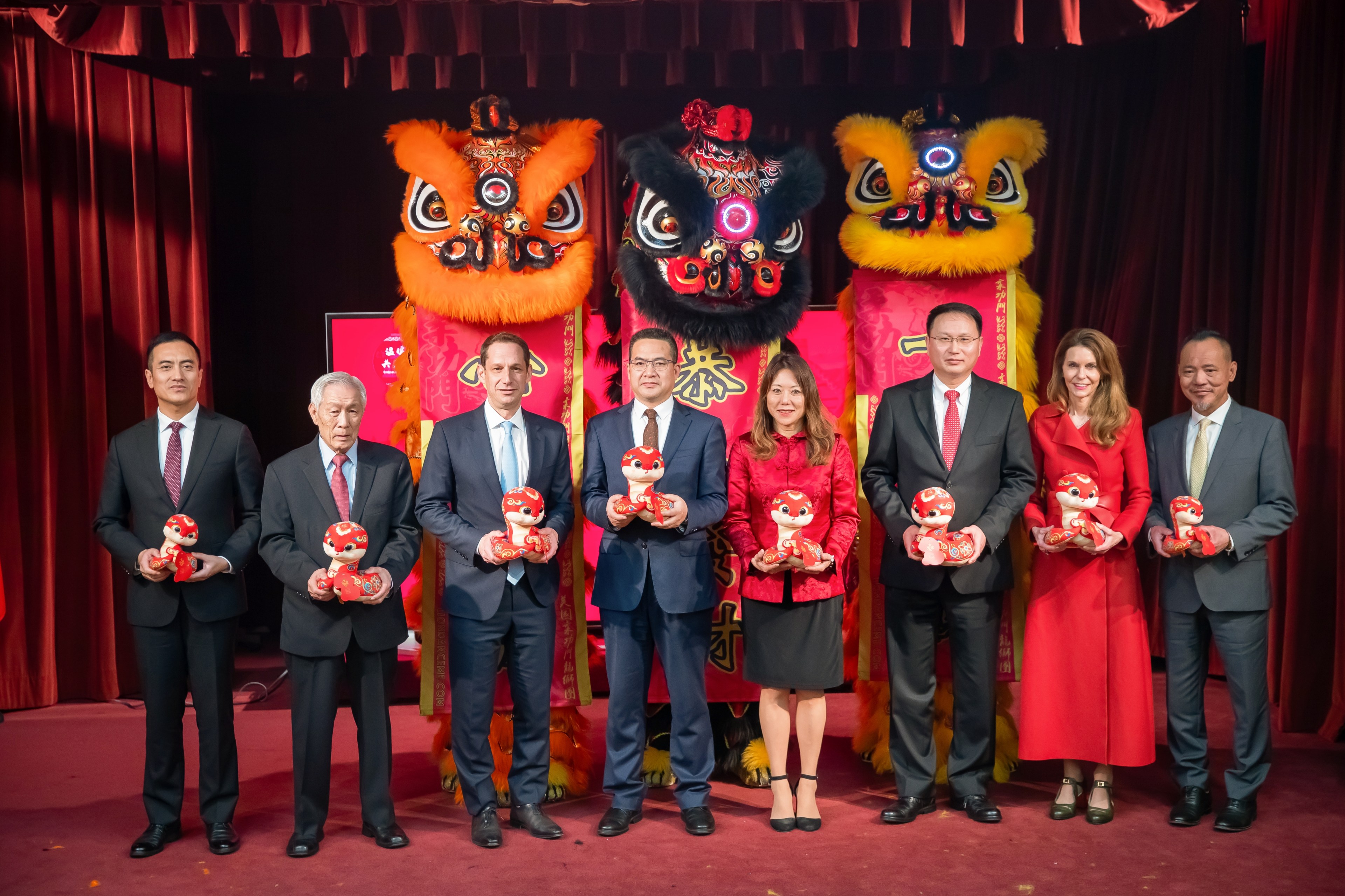 Eight people in formal attire stand on a stage holding red toys, with three large, colorful lion dance heads in the background and red curtains.