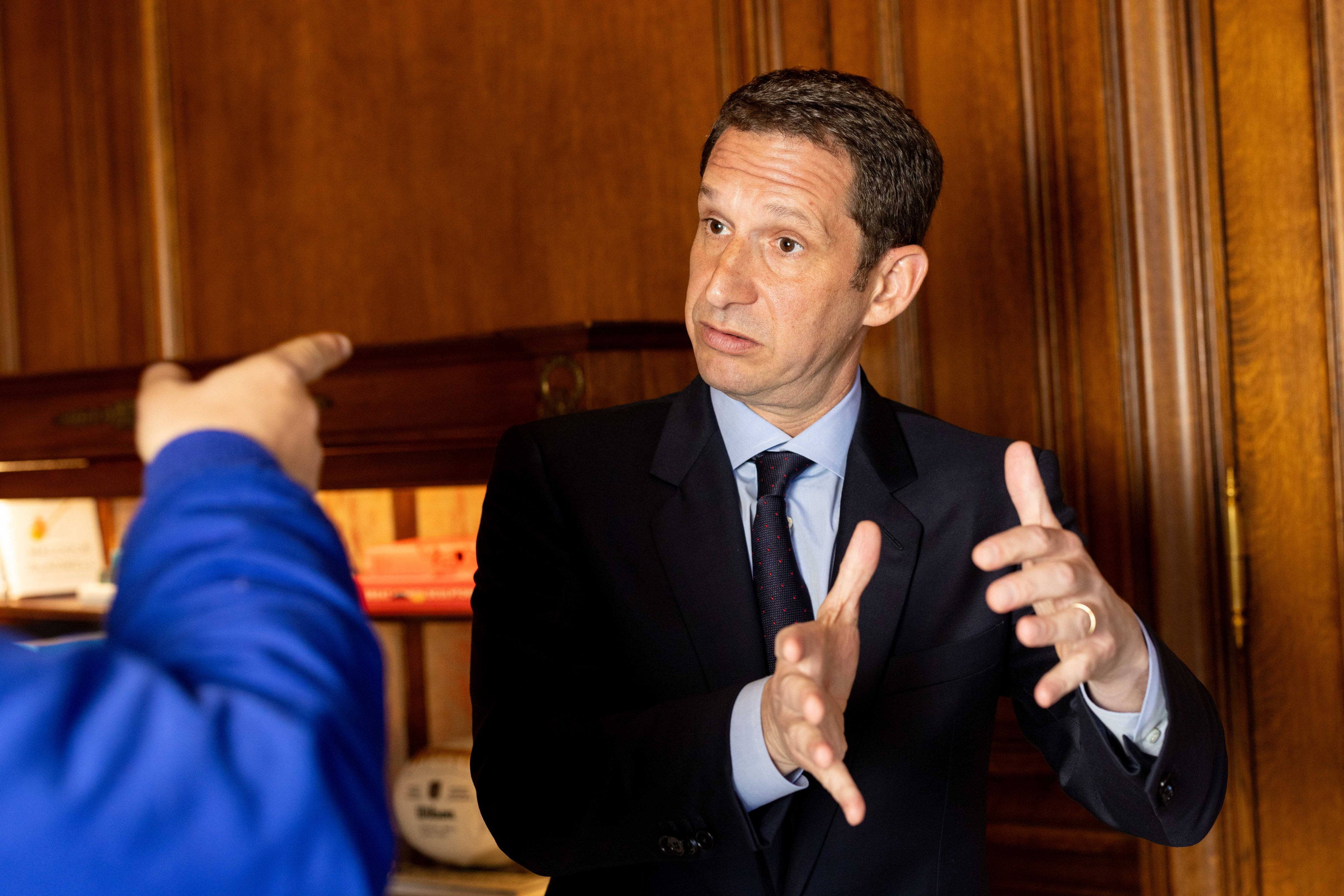 A man in a suit gestures expressively with his hands while speaking to someone in a blue jacket, inside a room with wooden walls and a shelf of items.