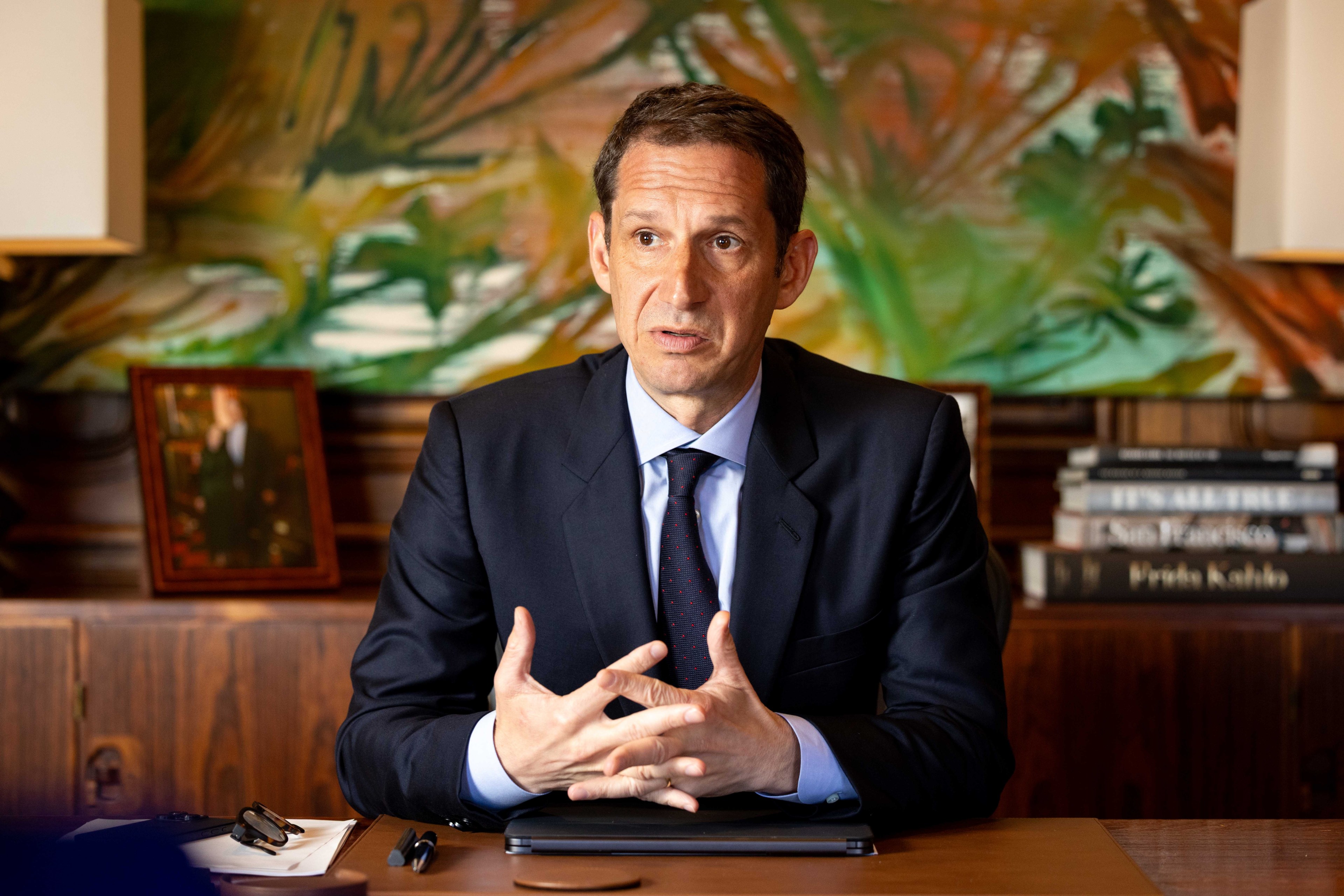 A man in a suit sits at a desk, hands clasped. Behind him are a colorful abstract painting, a framed photo, and a small stack of books.