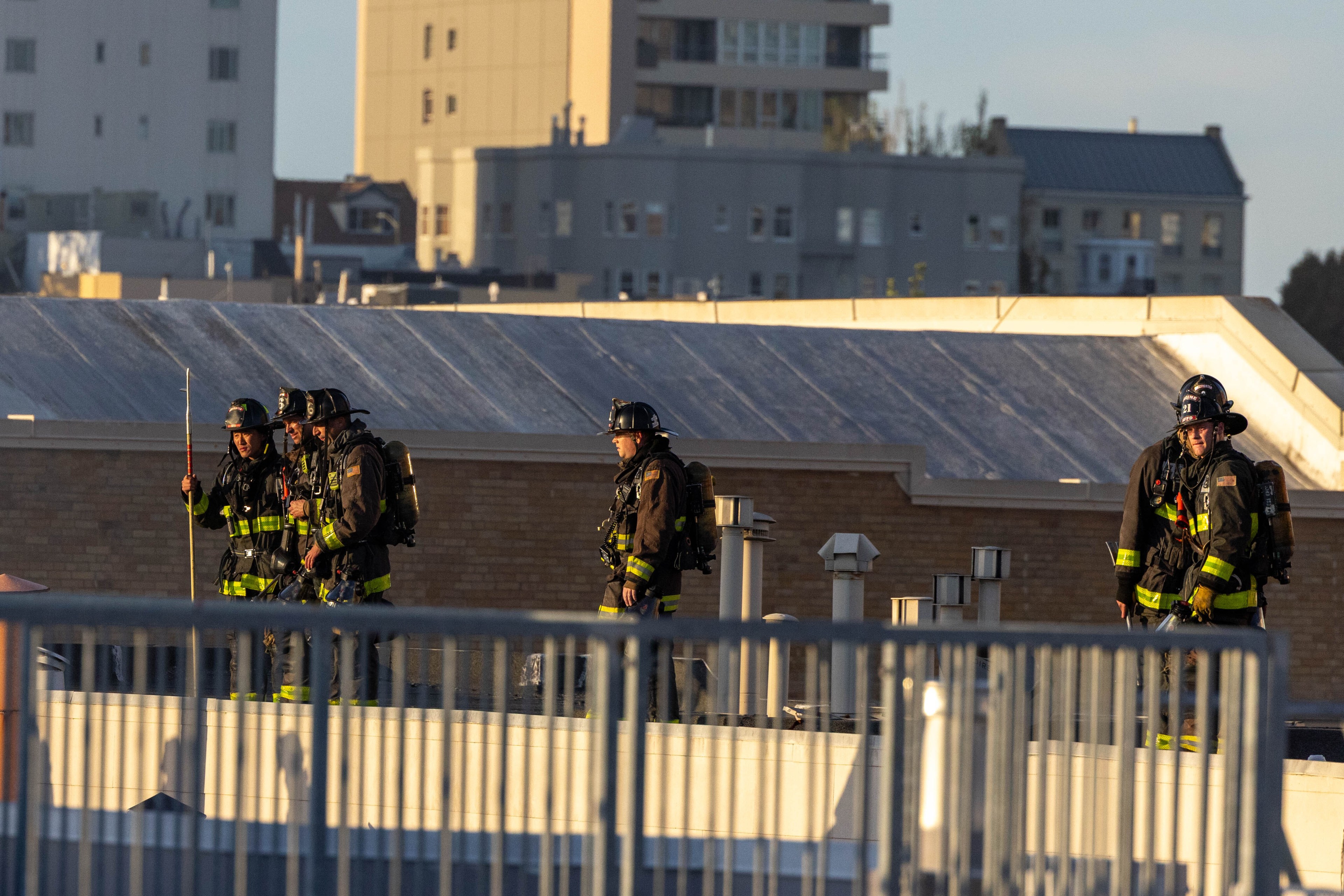 Four firefighters in full gear stand on a rooftop with urban buildings in the background, illuminated by sunlight.