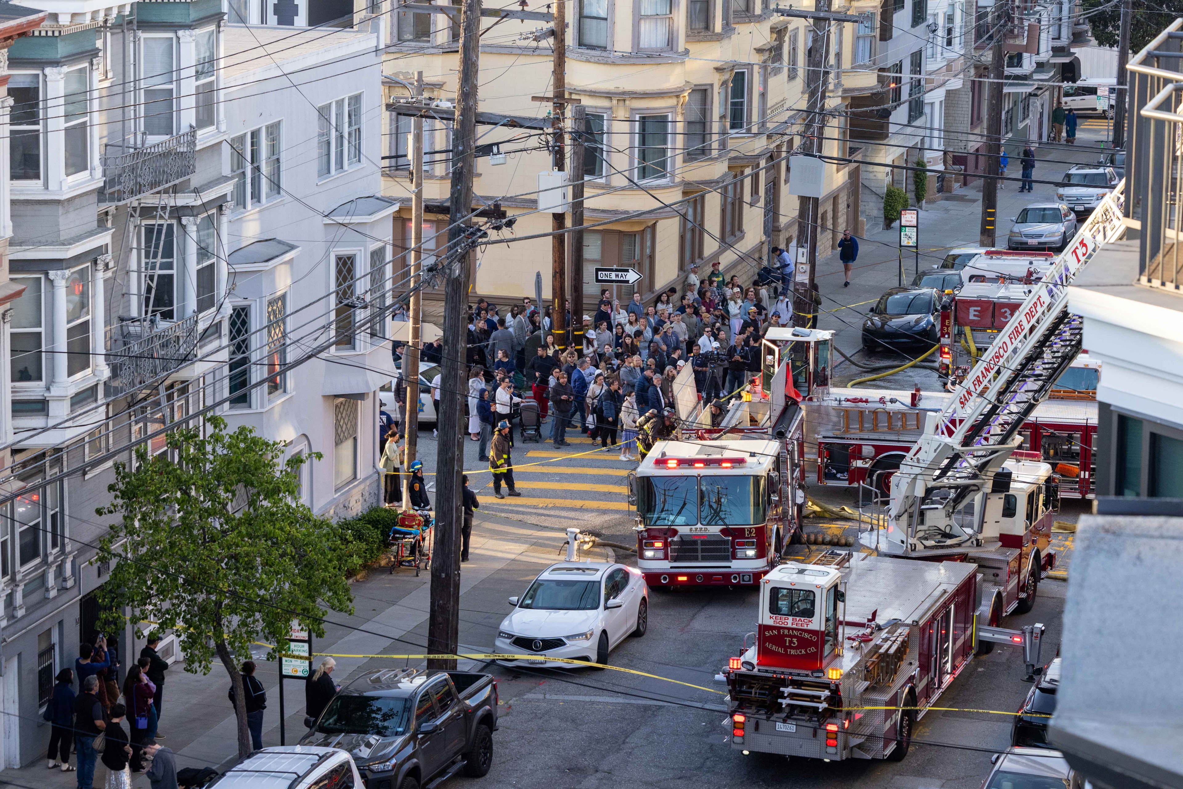 The image shows a crowded street with fire trucks, firemen, and people gathered on the sidewalk. Bright lights are seen, and yellow tape cordons off the area.