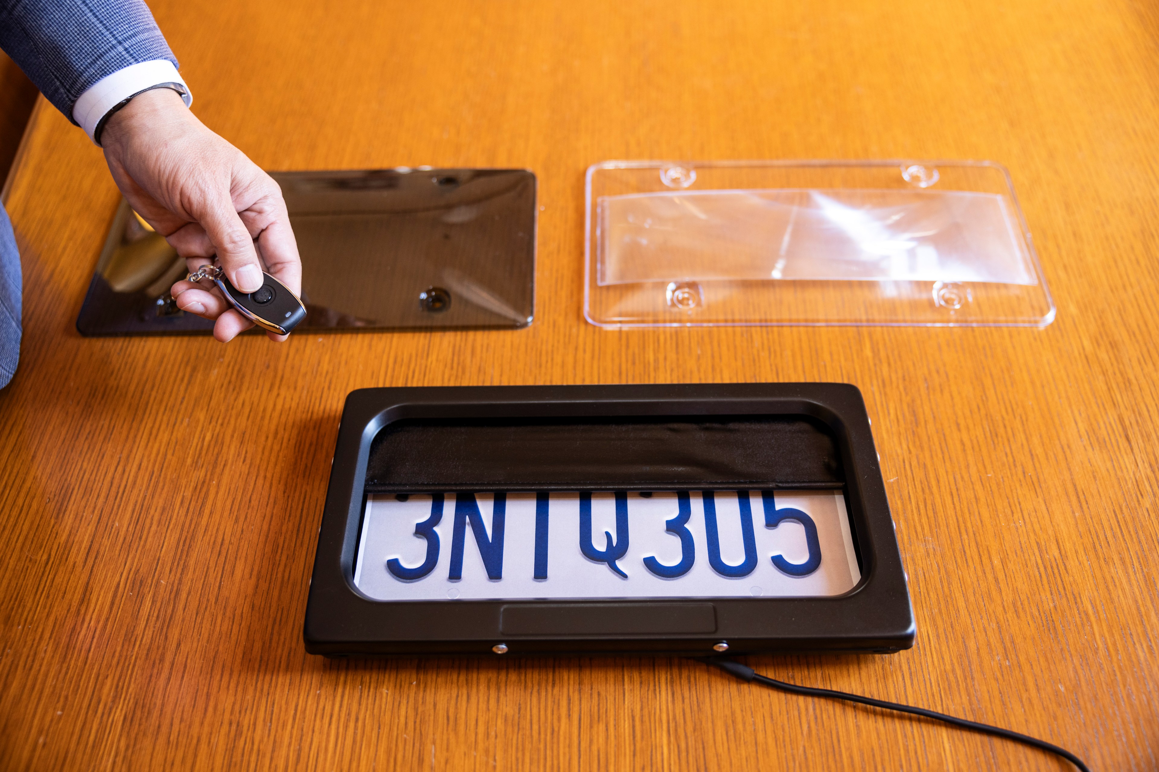 A hand holds a remote over a wooden table with a license plate device. Two plastic covers and a partially obscured plate with "3NTQ305" are visible.