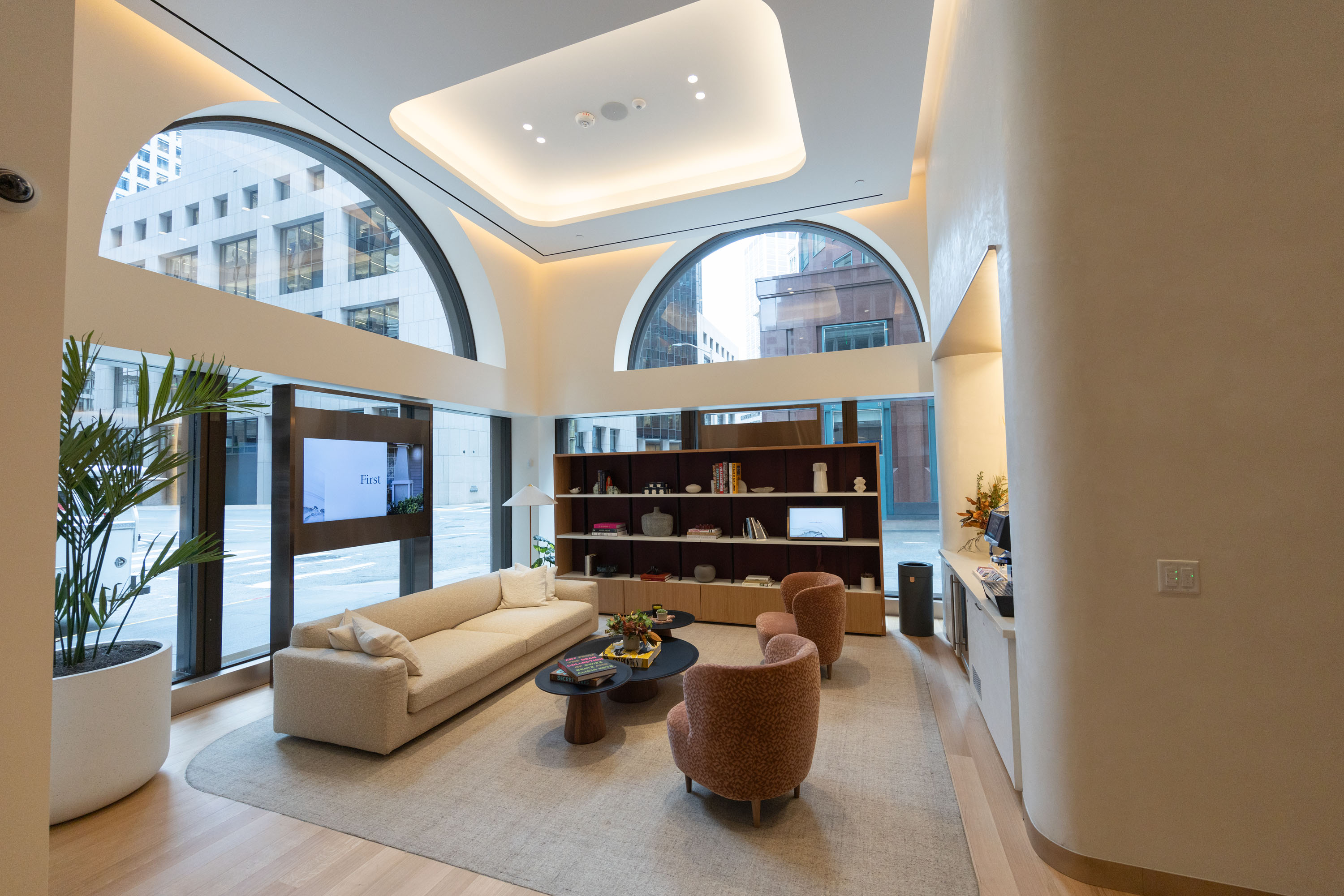 The image shows a modern, cozy lounge with large arched windows, soft lighting, plush seating, a beige couch, and two armchairs around a coffee table.
