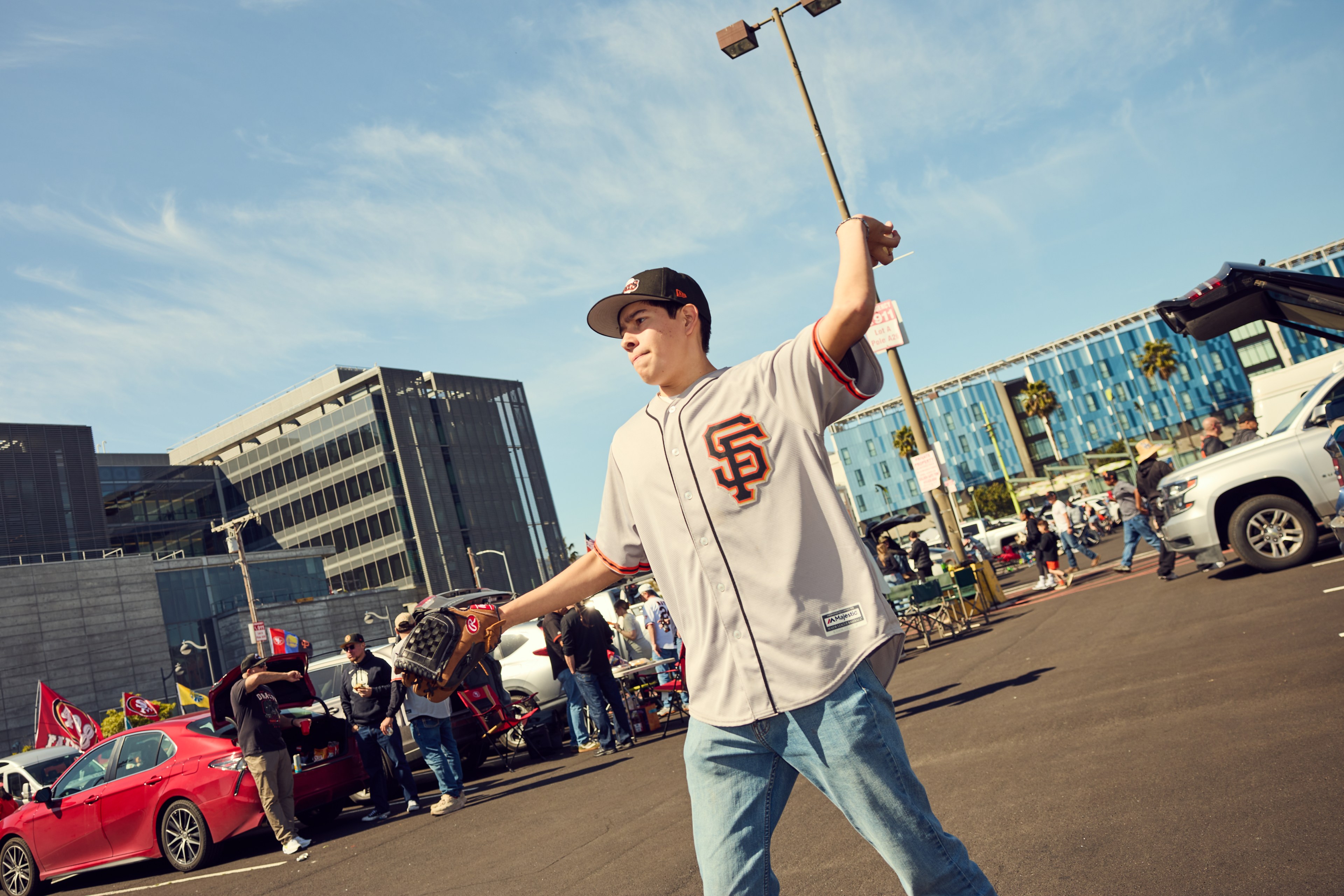 A person in a baseball jersey and cap is playfully throwing a baseball in a parking lot, with cars and a few people in the background.