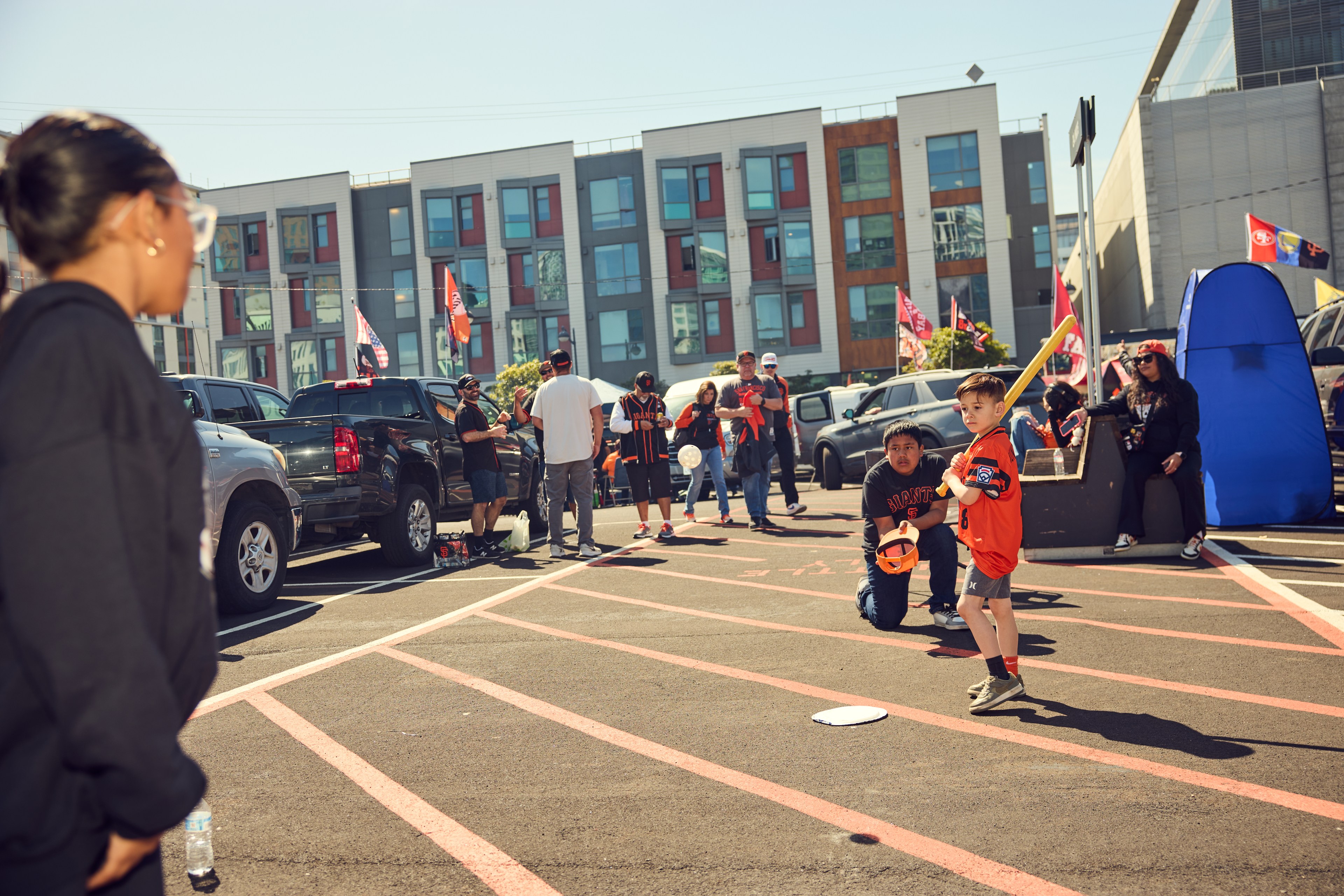 A child with a yellow bat stands ready to hit a ball in a lively parking lot, surrounded by people in sports jerseys and flags, indicating a festive atmosphere.