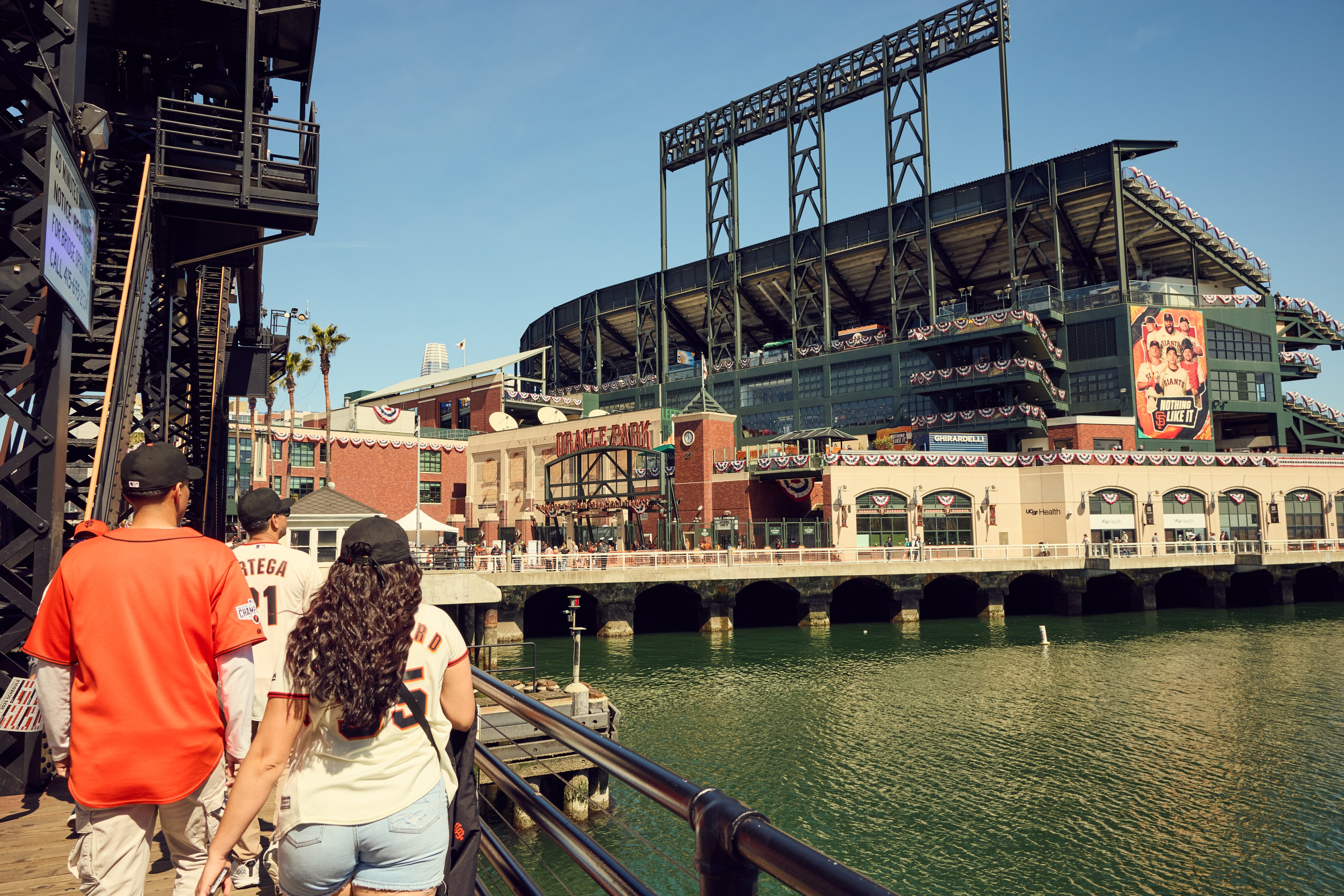 A group of people in baseball jerseys walk on a bridge towards a large stadium adorned with banners. The water is visible below, and the sky is clear.
