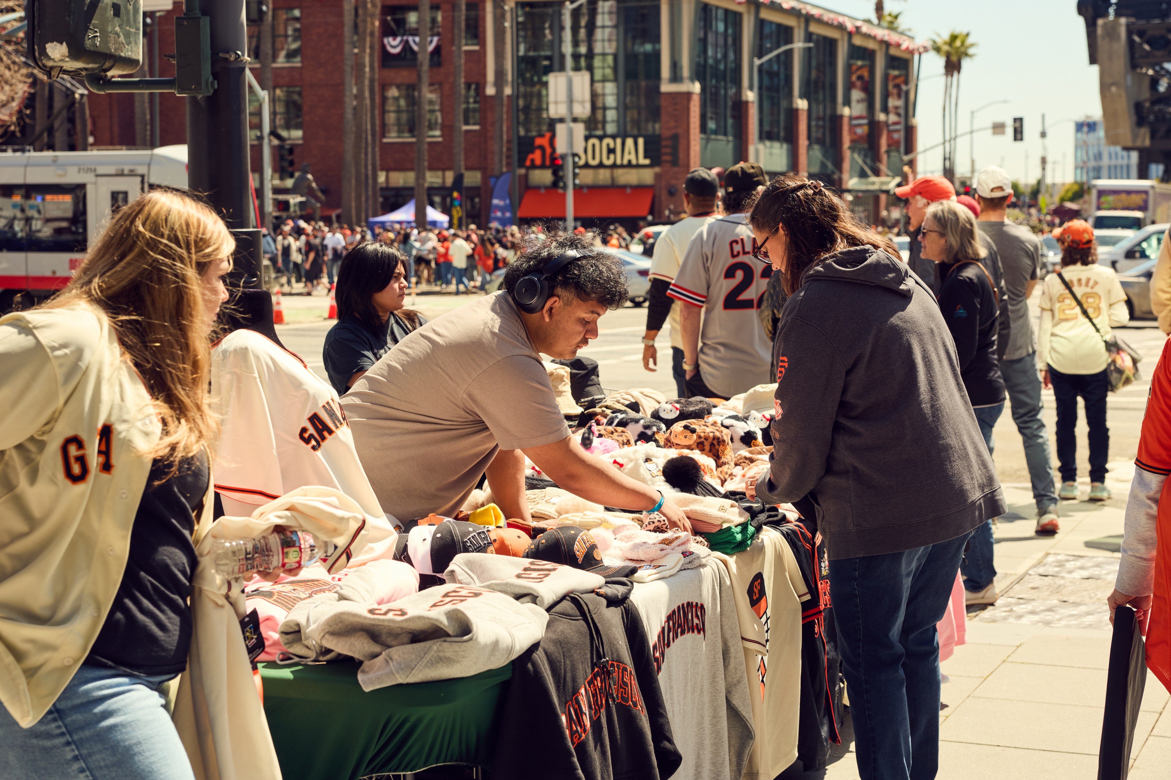 People are browsing a street vendor's table full of San Francisco Giants merchandise, including shirts, caps, and plush toys, near a stadium.