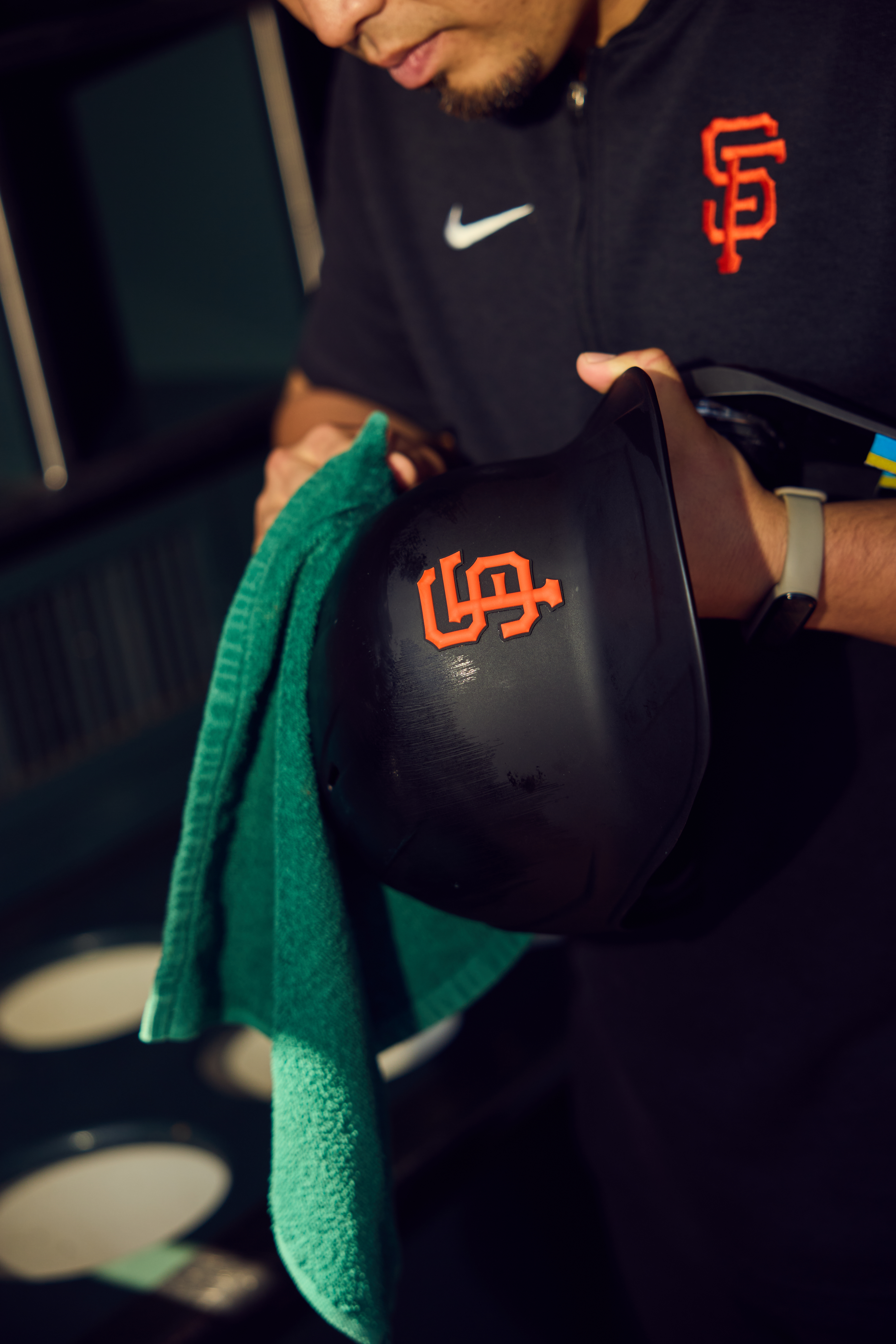 A person cleans a black helmet with an orange logo using a green cloth. The individual wears a dark top with a similar orange logo and a smartwatch.