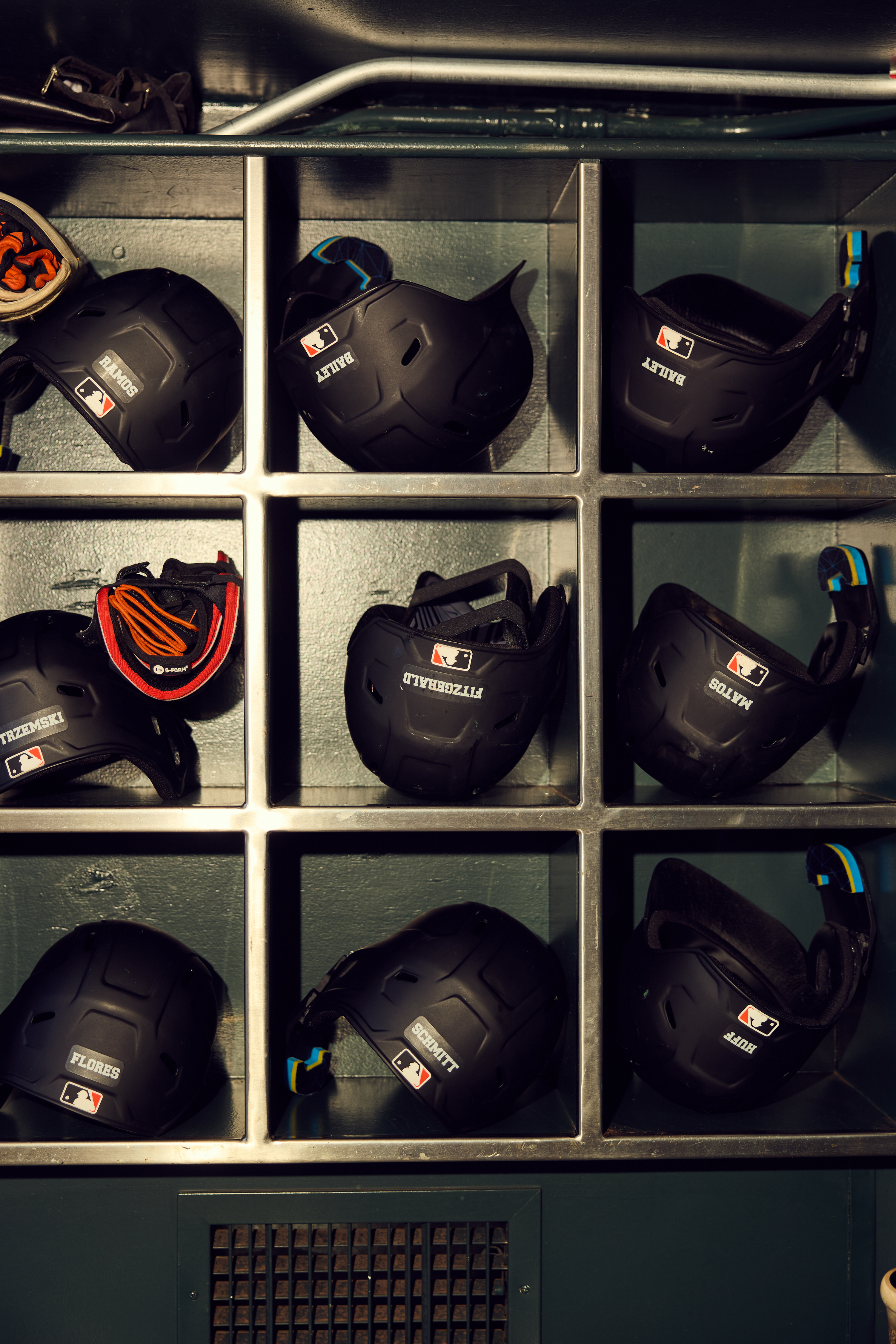 The image shows a metal shelf with nine compartments holding black baseball helmets. Each compartment has a helmet with a name and the MLB logo on it.
